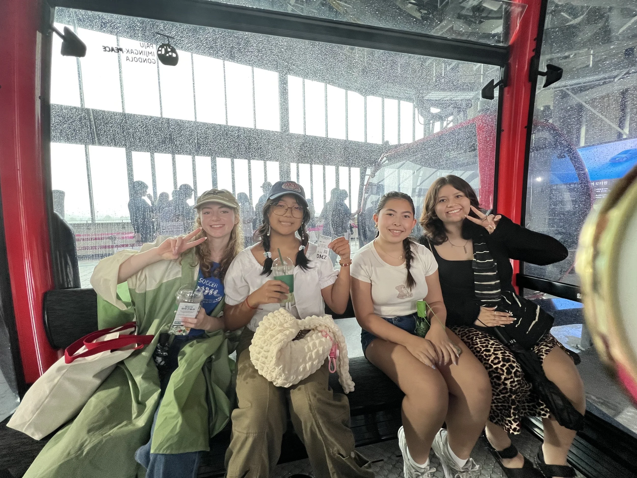 Four young women sitting inside a cable car, smiling and making peace signs. They have casual clothing and are holding drinks and bags.