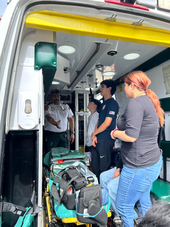 People inside an ambulance, including a paramedic explaining something to a woman and young individuals, with medical equipment visible.