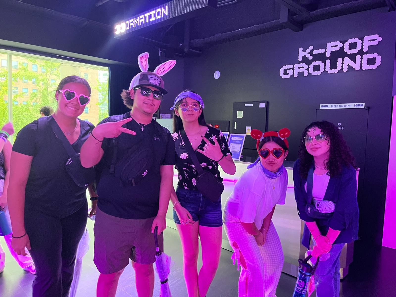 Group of five people wearing playful accessories and sunglasses, standing inside a K-pop themed arcade or entertainment venue with vibrant lighting.