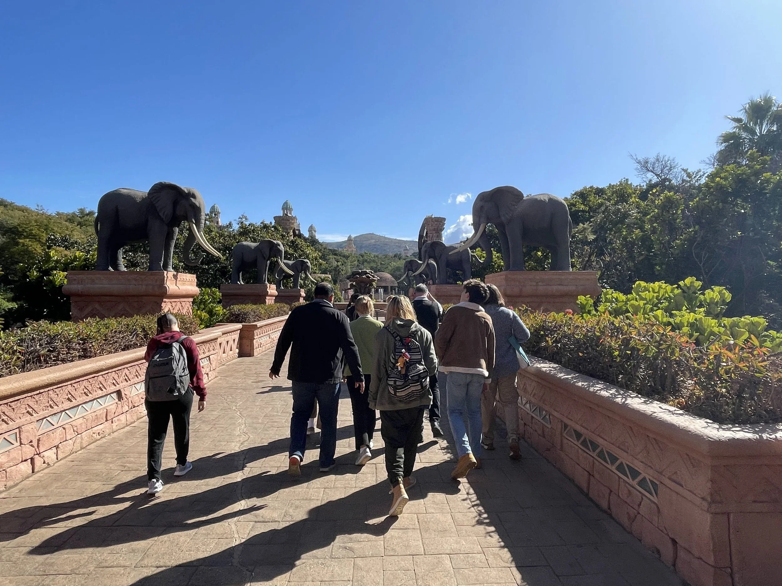 Tourists walking along a pathway lined with elephant statues in an outdoor park with lush greenery and blue sky.