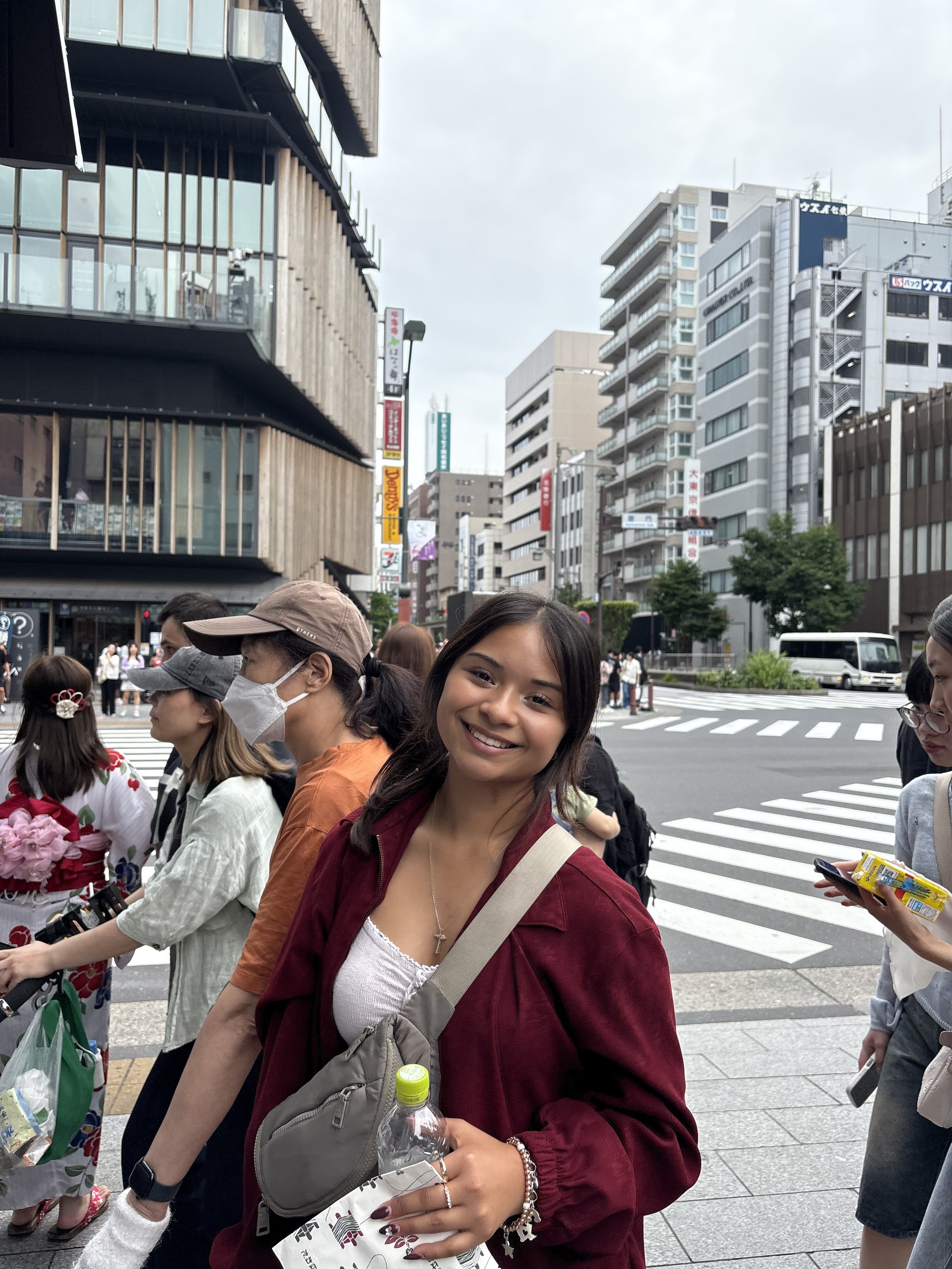 Smiling young woman with a beige crossbody bag containing a water bottle, standing on a busy city street with pedestrians, tall buildings, and crosswalks visible in the background.