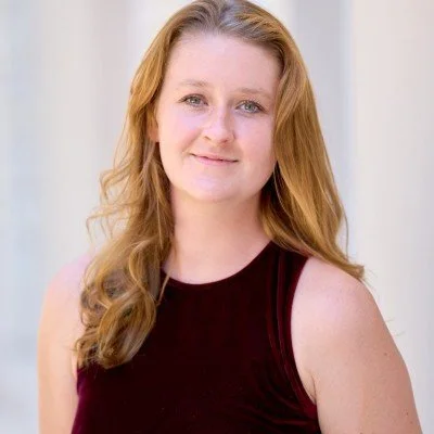Portrait of a young woman with long, wavy red hair, wearing a sleeveless maroon top, smiling softly, with a light-colored background.