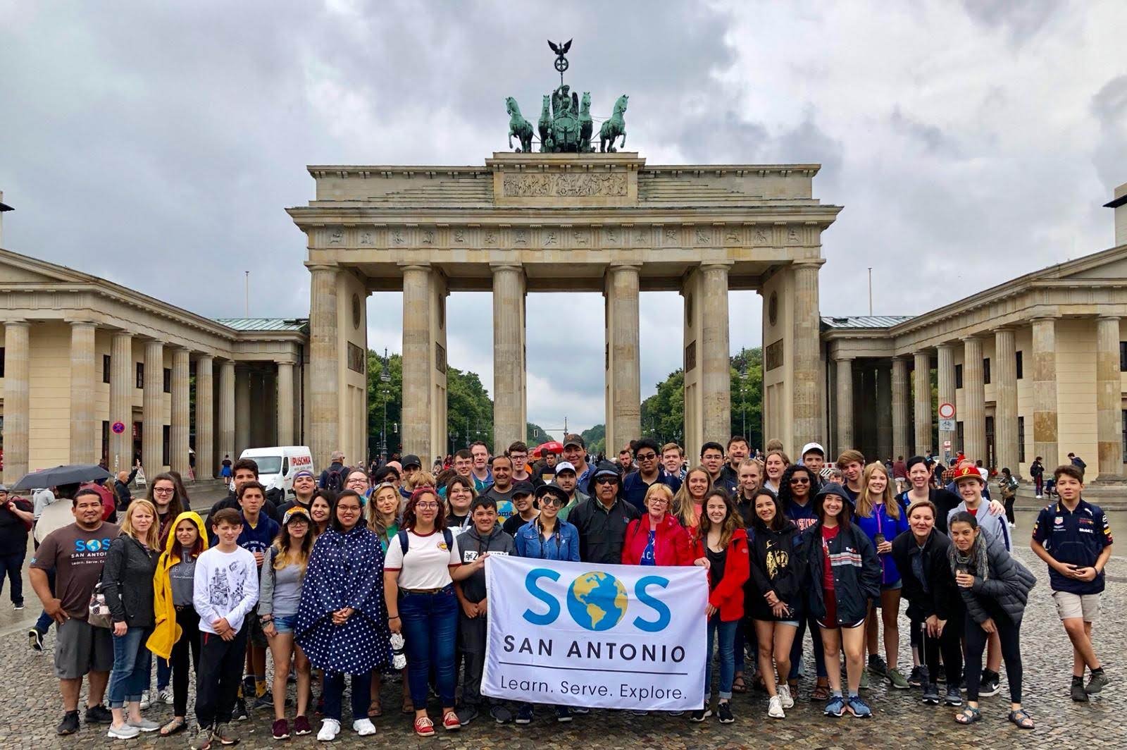 A large group of people holding a SOS San Antonio banner posing in front of the Brandenburg Gate in Berlin, Germany during an overcast day.