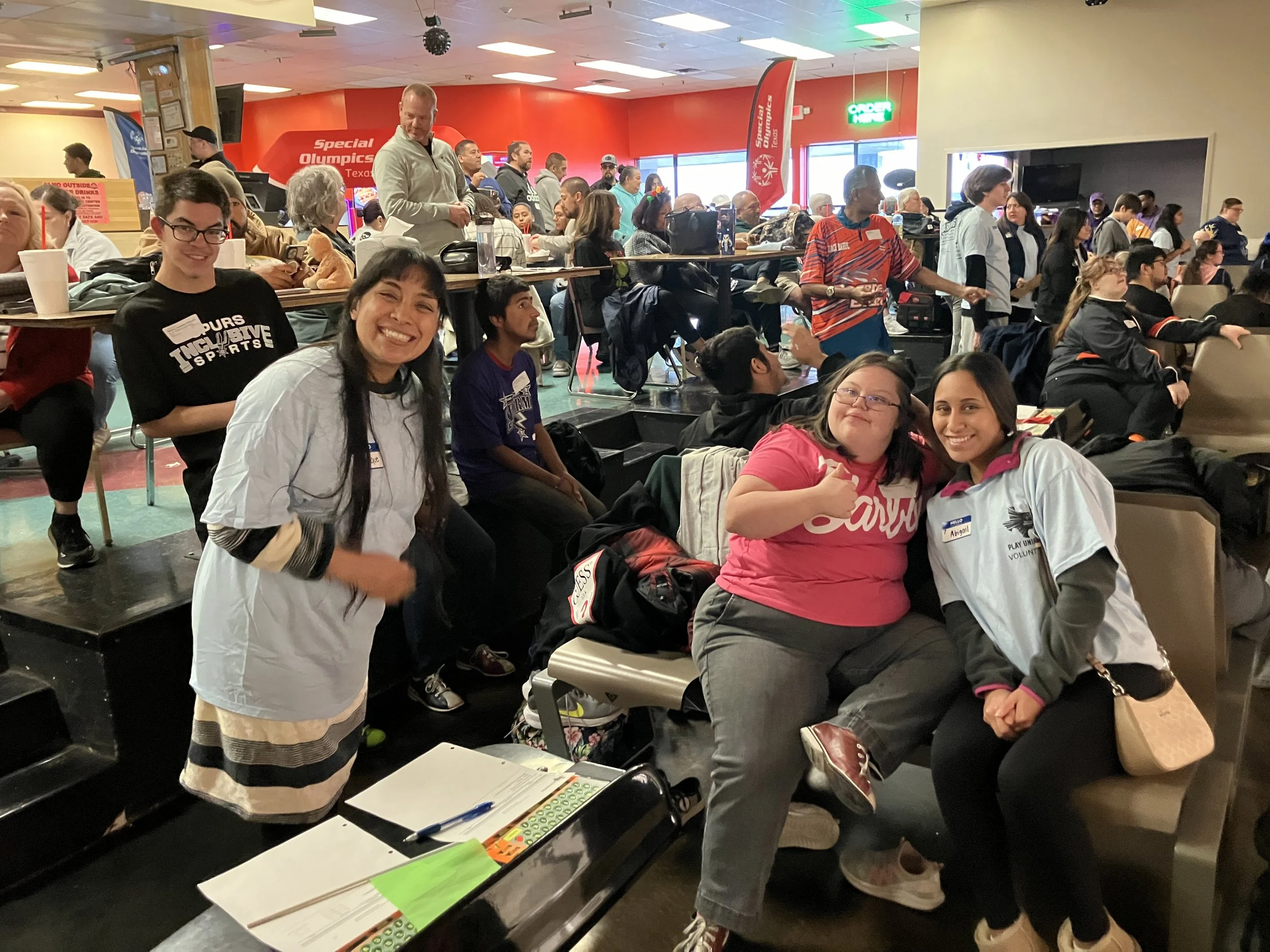 Group of people at an indoor event, some sitting and some standing, with tables, chairs, and banners in the background.