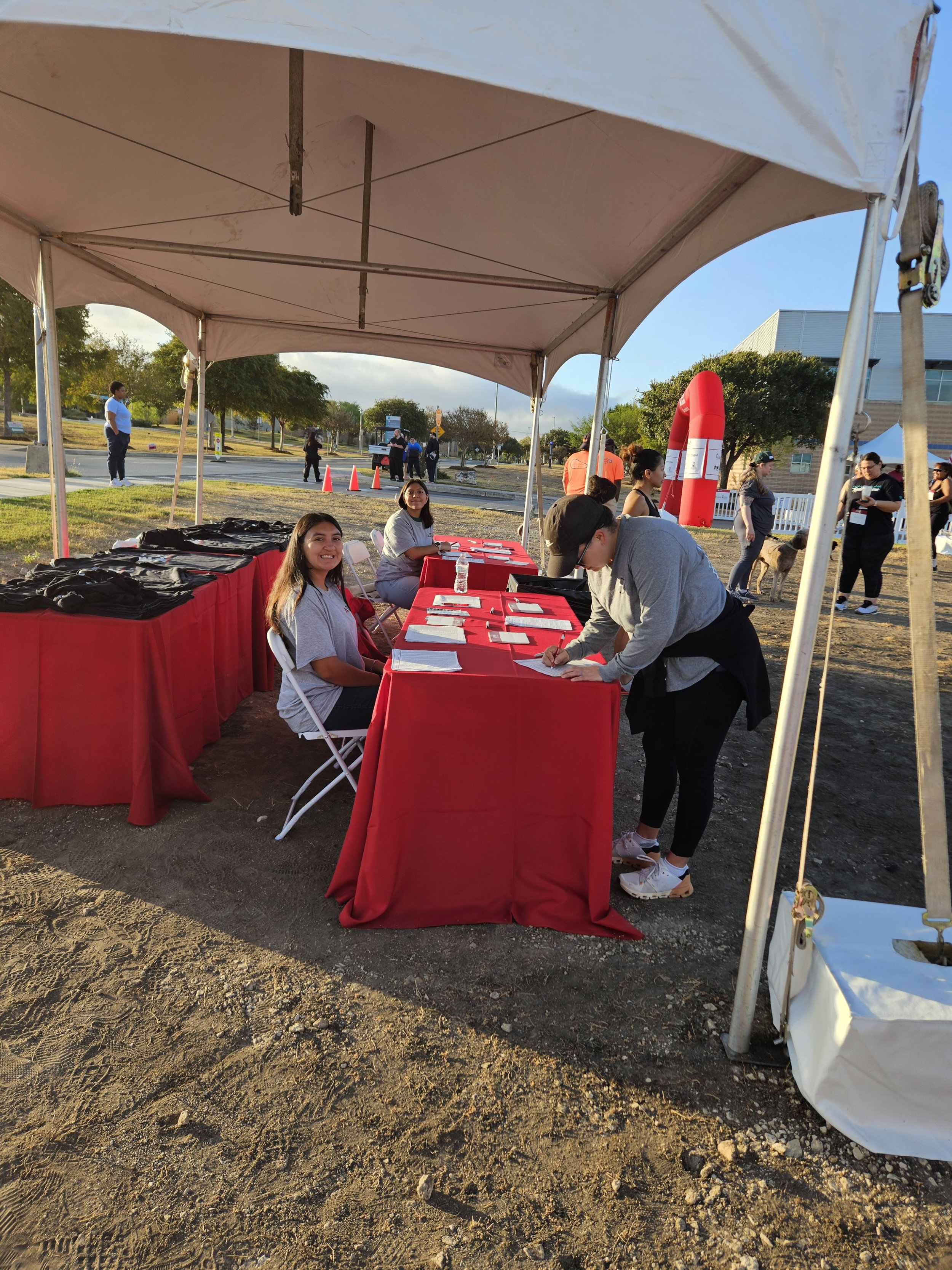People sitting and standing at a registration tent with red tablecloths at an outdoor event, with several people walking in the background, traffic cones, and a large red inflatable object.