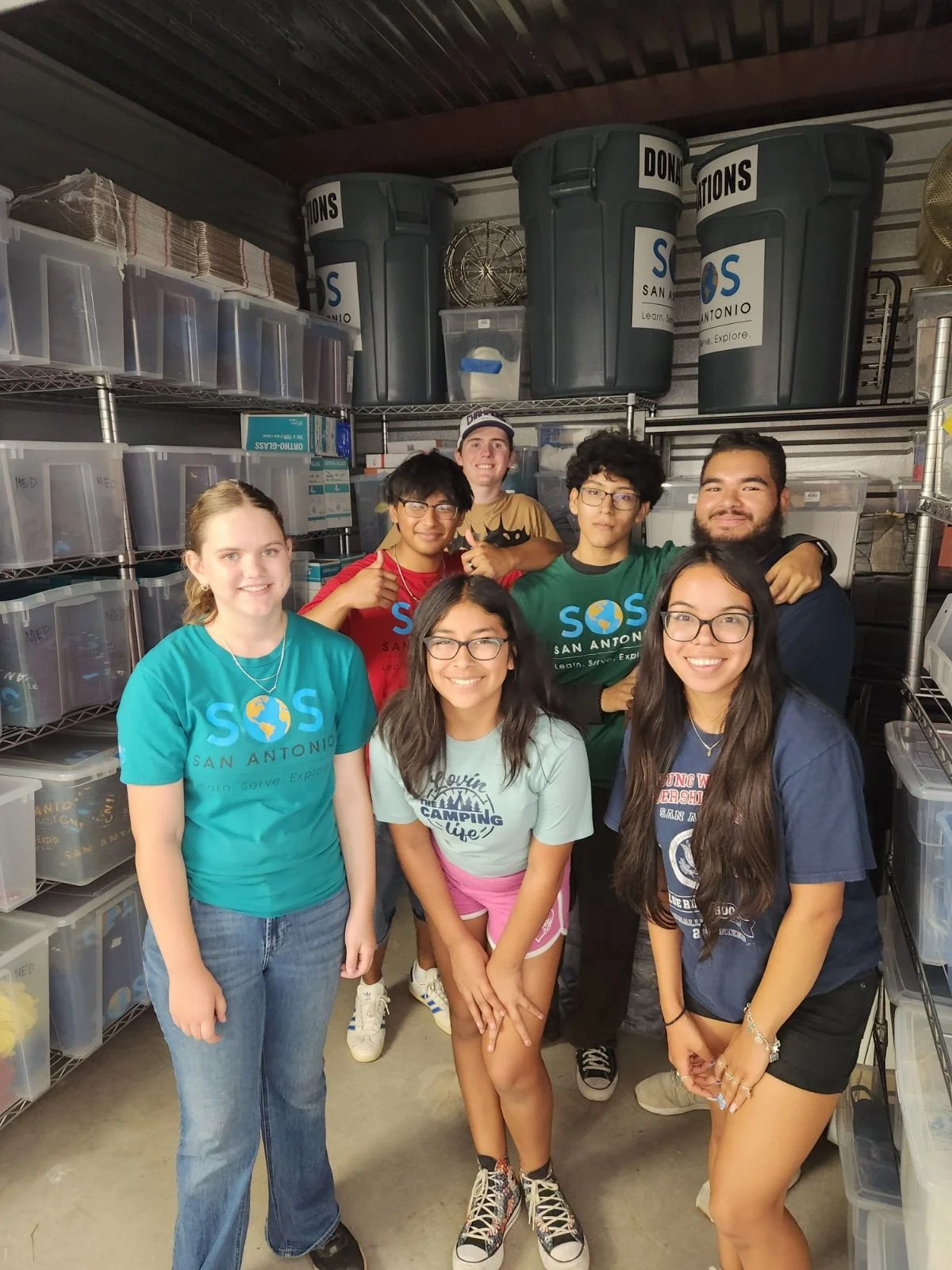 Group of seven young people posing in a storage room with plastic bins and large trash cans labeled 'Donations', all smiling.
