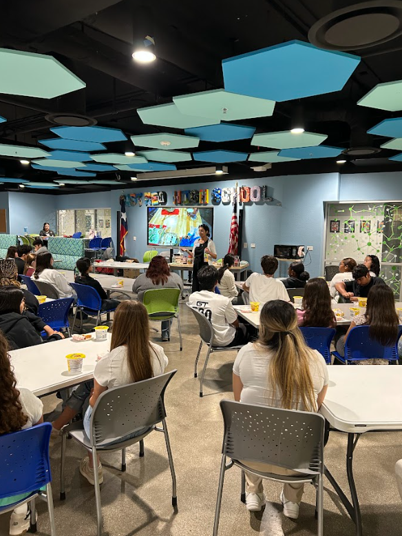 A classroom with students seated at tables, a teacher standing at the front, and a colorful digital screen. The room has blue and green ceiling panels and is decorated with a banner saying 'Owen J. Roberts School' and flags.