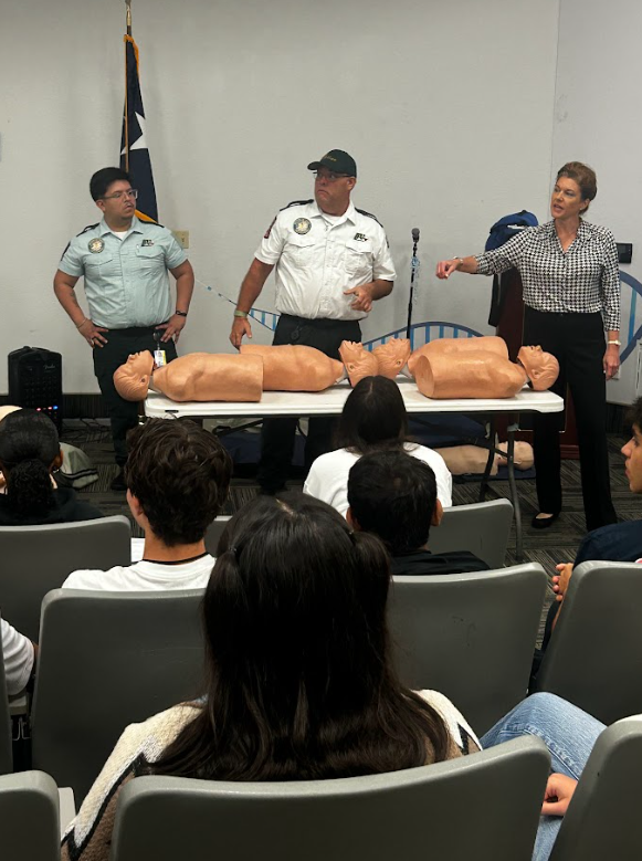 Three instructors, a man in a white security uniform, a woman in a checkered shirt, and a man in a light blue shirt, stand in front of an audience of students seated in a classroom. They are demonstrating CPR techniques using adult mannequins on tabl