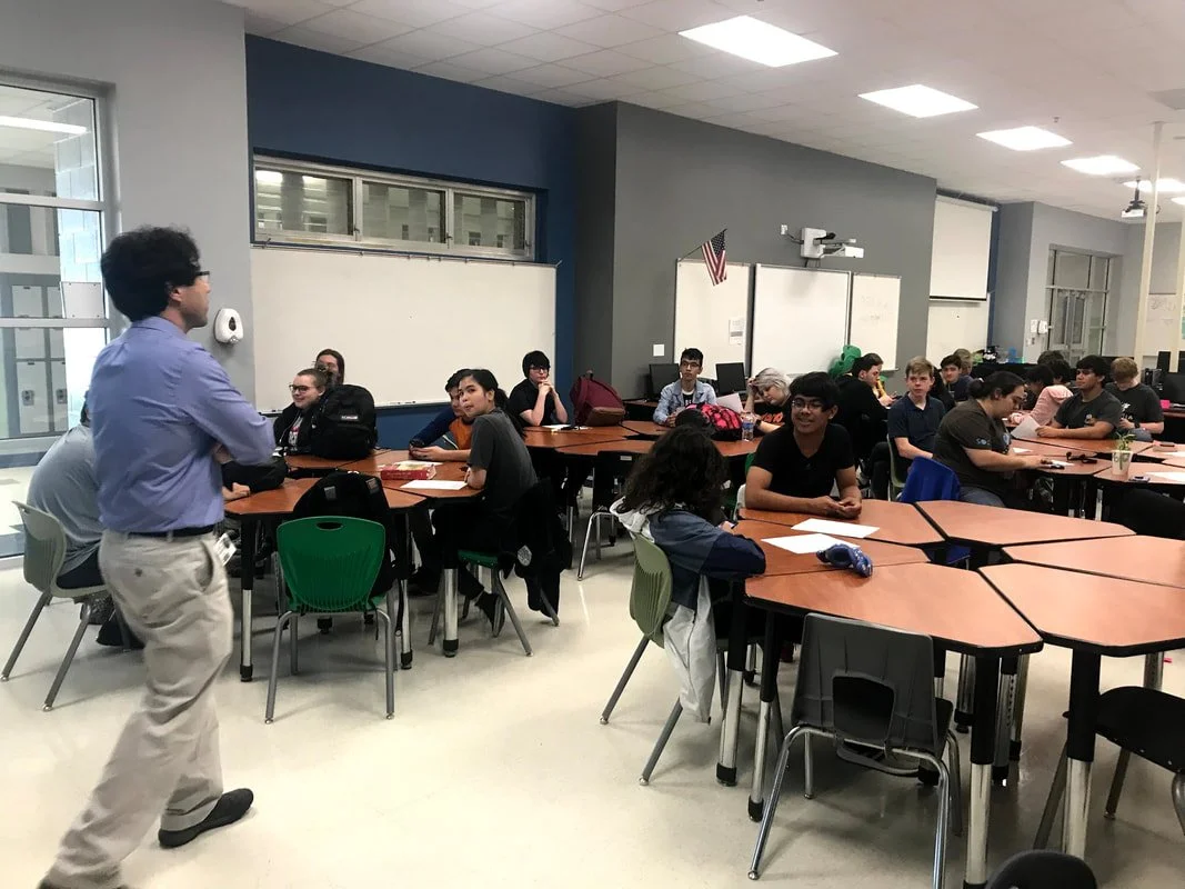 A classroom with students seated at tables listening to a teacher standing in front.