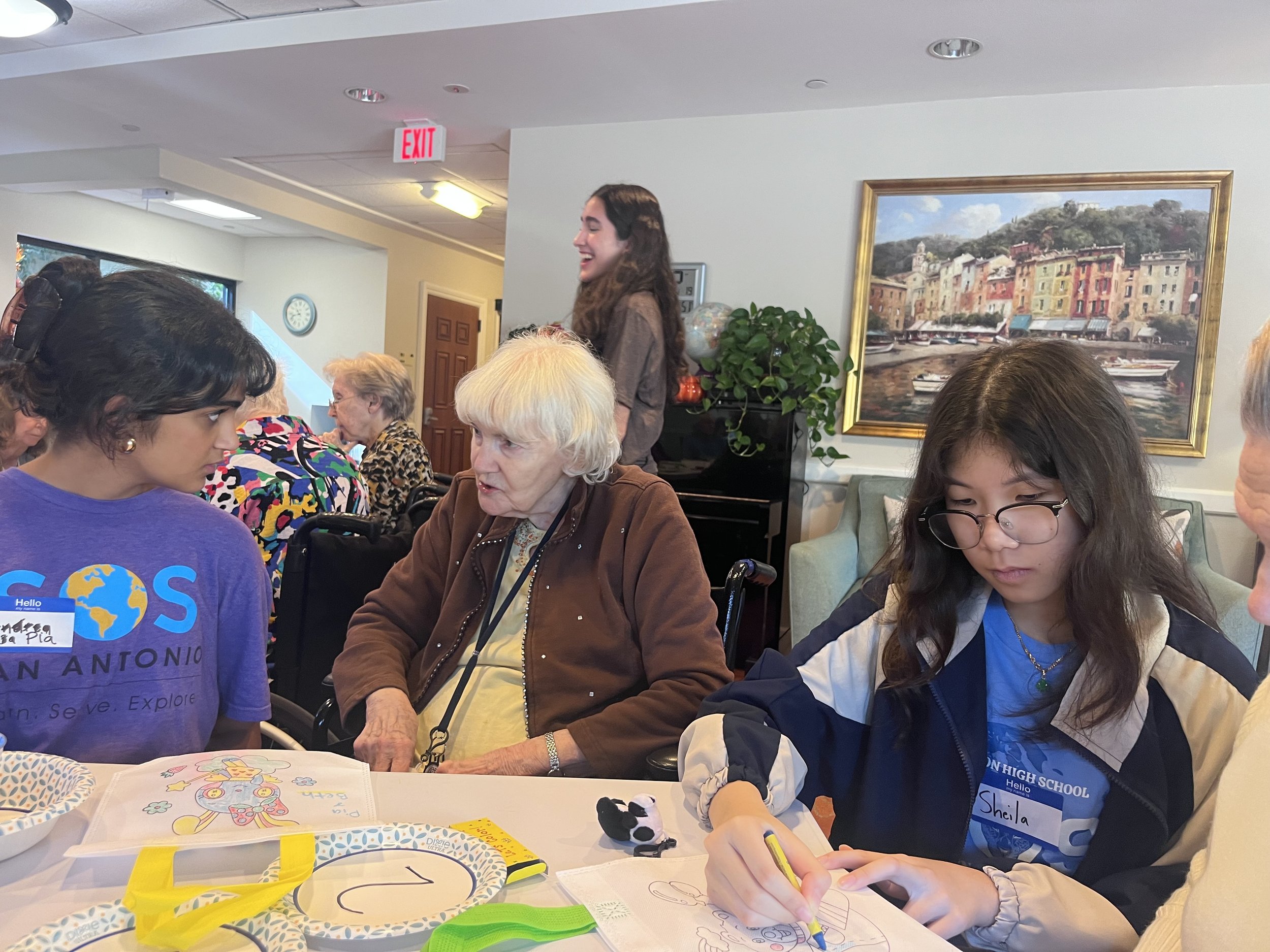 A group of people sitting at a table, engaging in conversation and drawing, with a young woman and an elderly woman in focus.