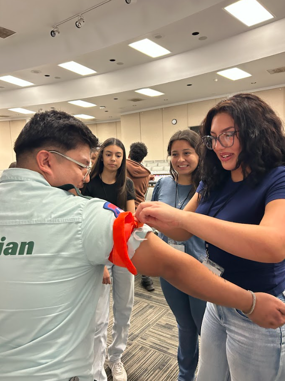 A group of young people in an indoor room, with one person in a light-colored uniform, is being helped with a red band as part of a medical training activity, while others observe.