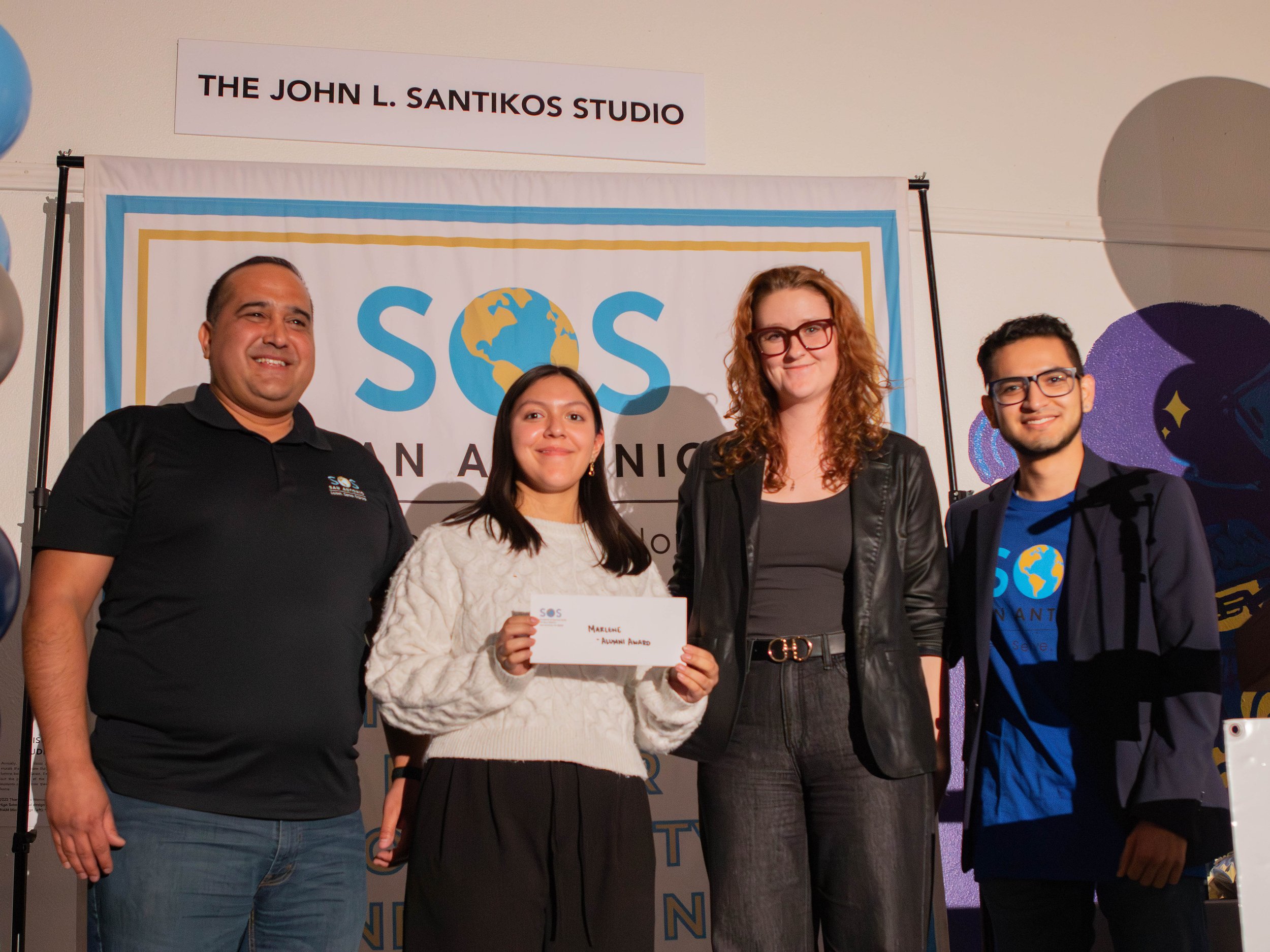 Group of four people standing in front of a banner at an award ceremony. One woman is holding an award certificate. All are smiling and dressed casually and semi-formally.