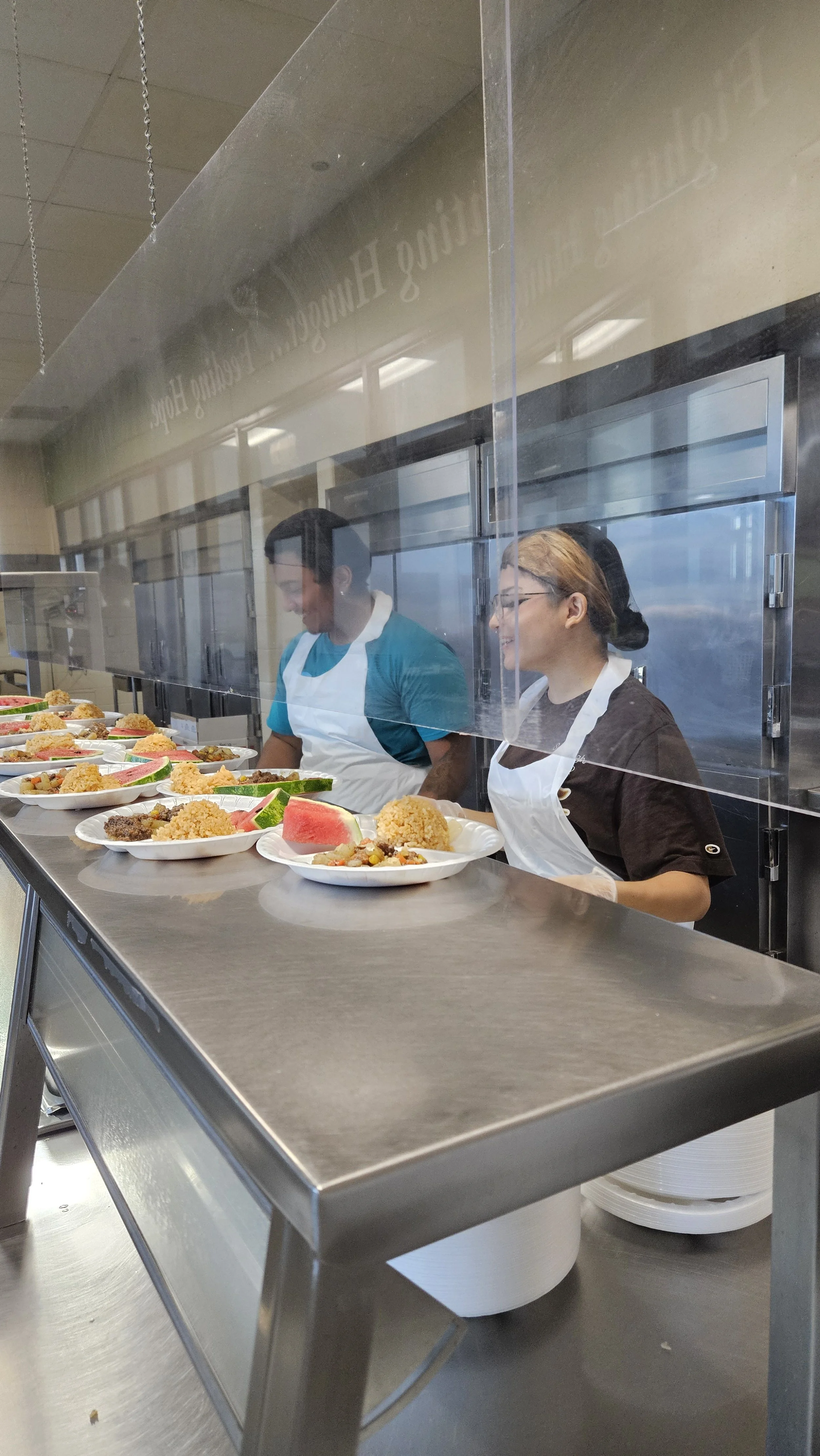 Two food service workers behind a glass divider preparing plates of food for serving in a cafeteria or dining hall.