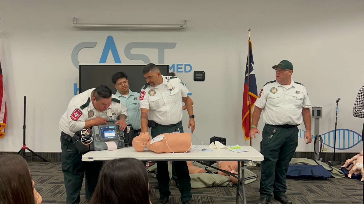 Medical professionals conducting a CPR training demonstration with a mannequin in a classroom setting, with American flags and a sign in the background.