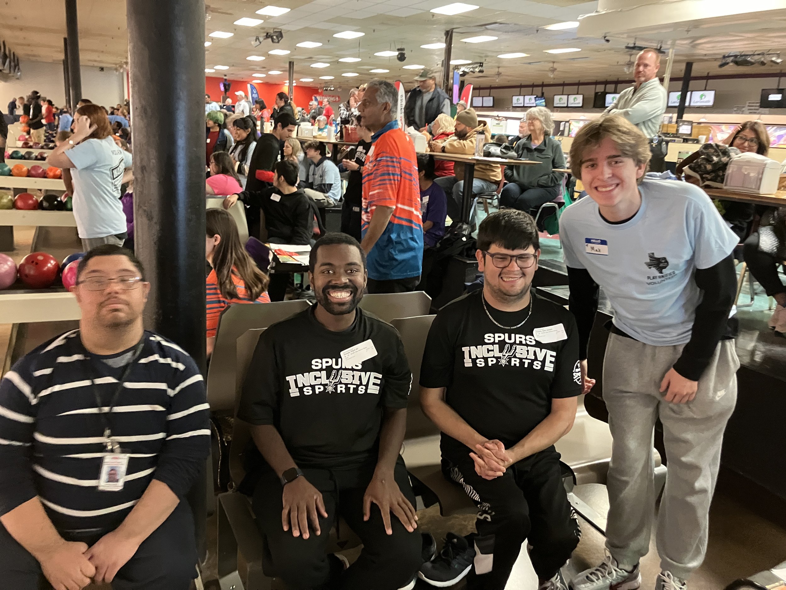 Four men sitting and standing in a bowling alley, smiling, with bowling balls and other players in the background.