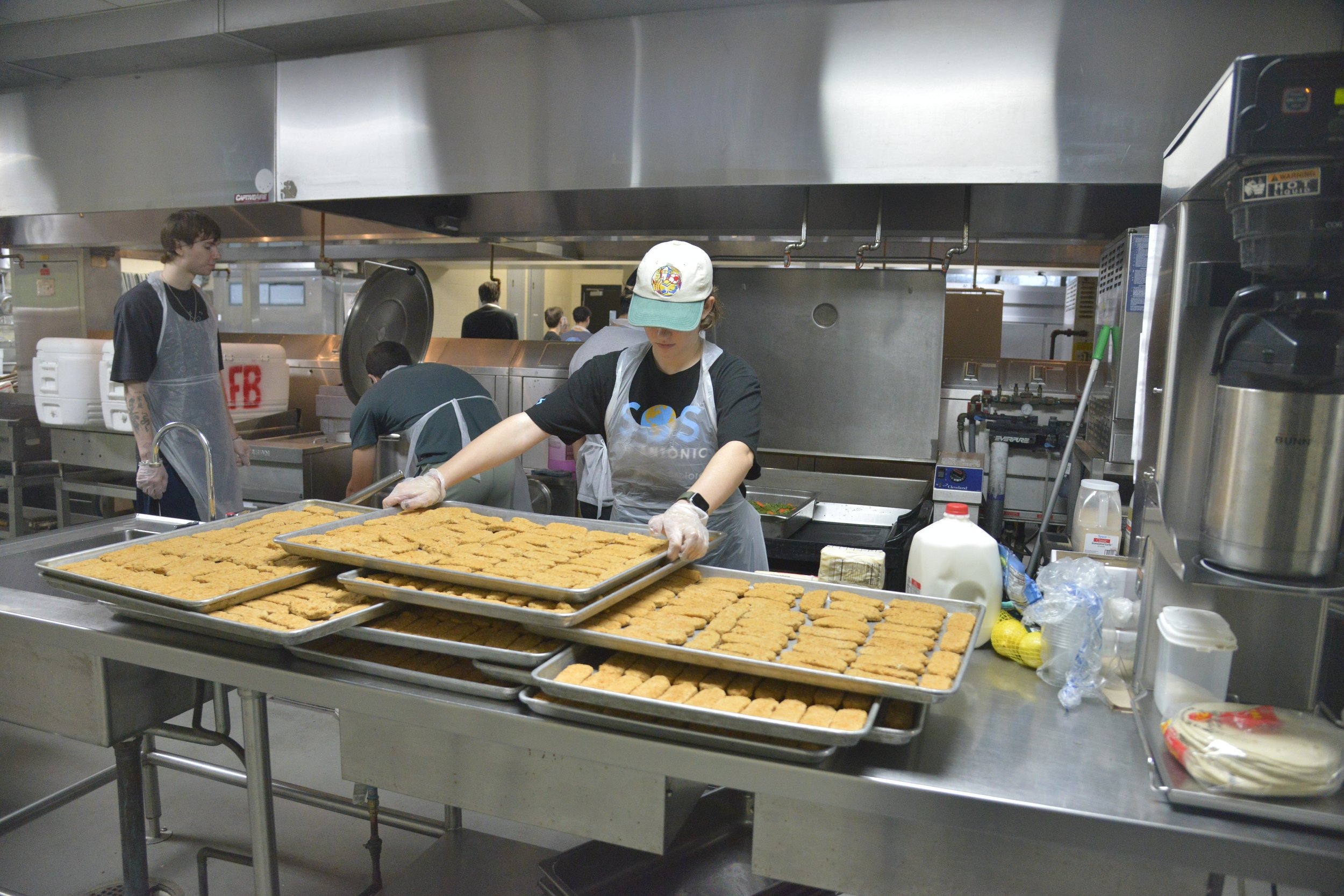 A person working in a commercial kitchen prepares trays of food, with other staff working in the background to be distributed. 