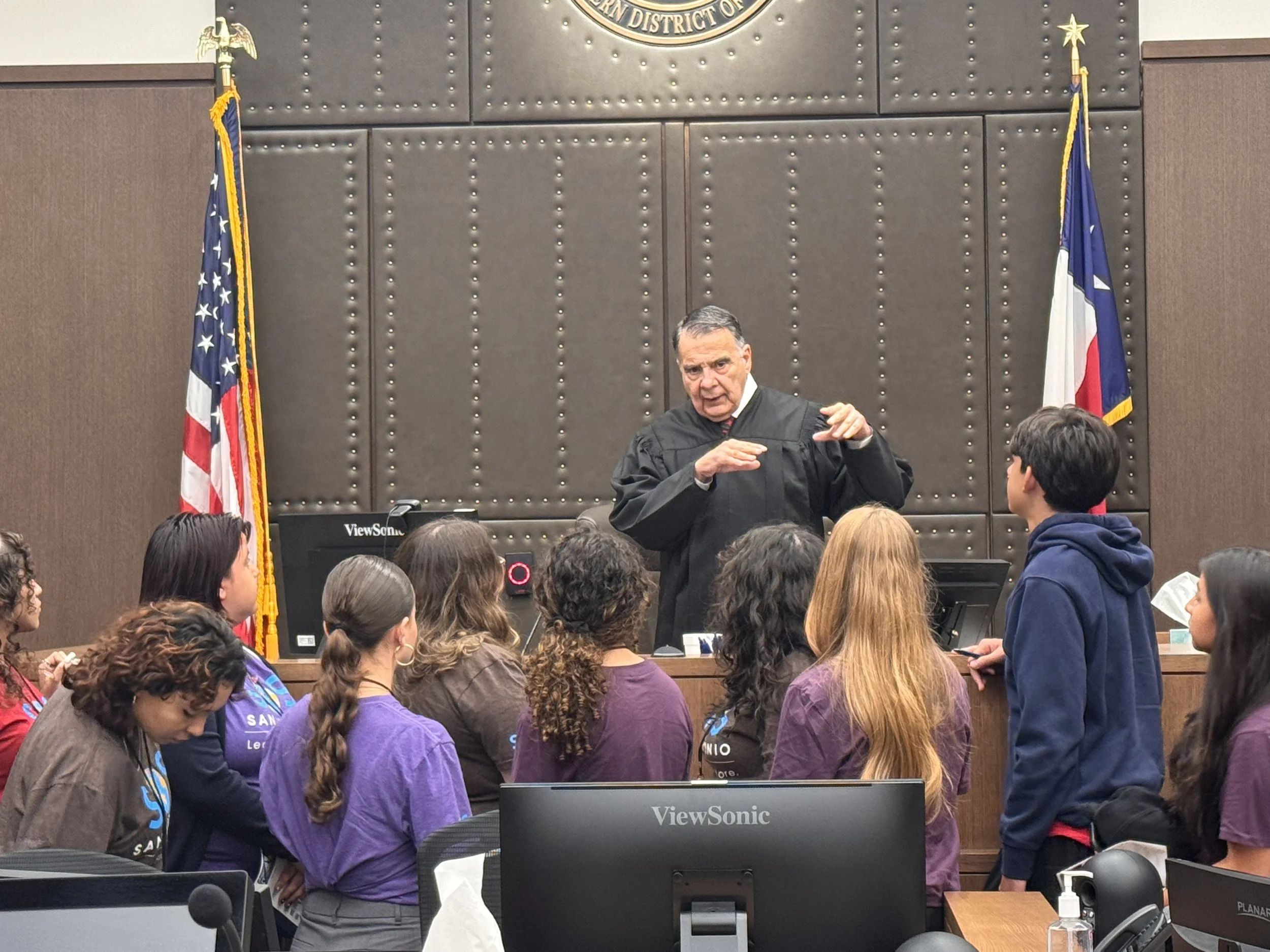 Judge in black robe speaking to a group of students in a courtroom with American and Texas flags in the background.