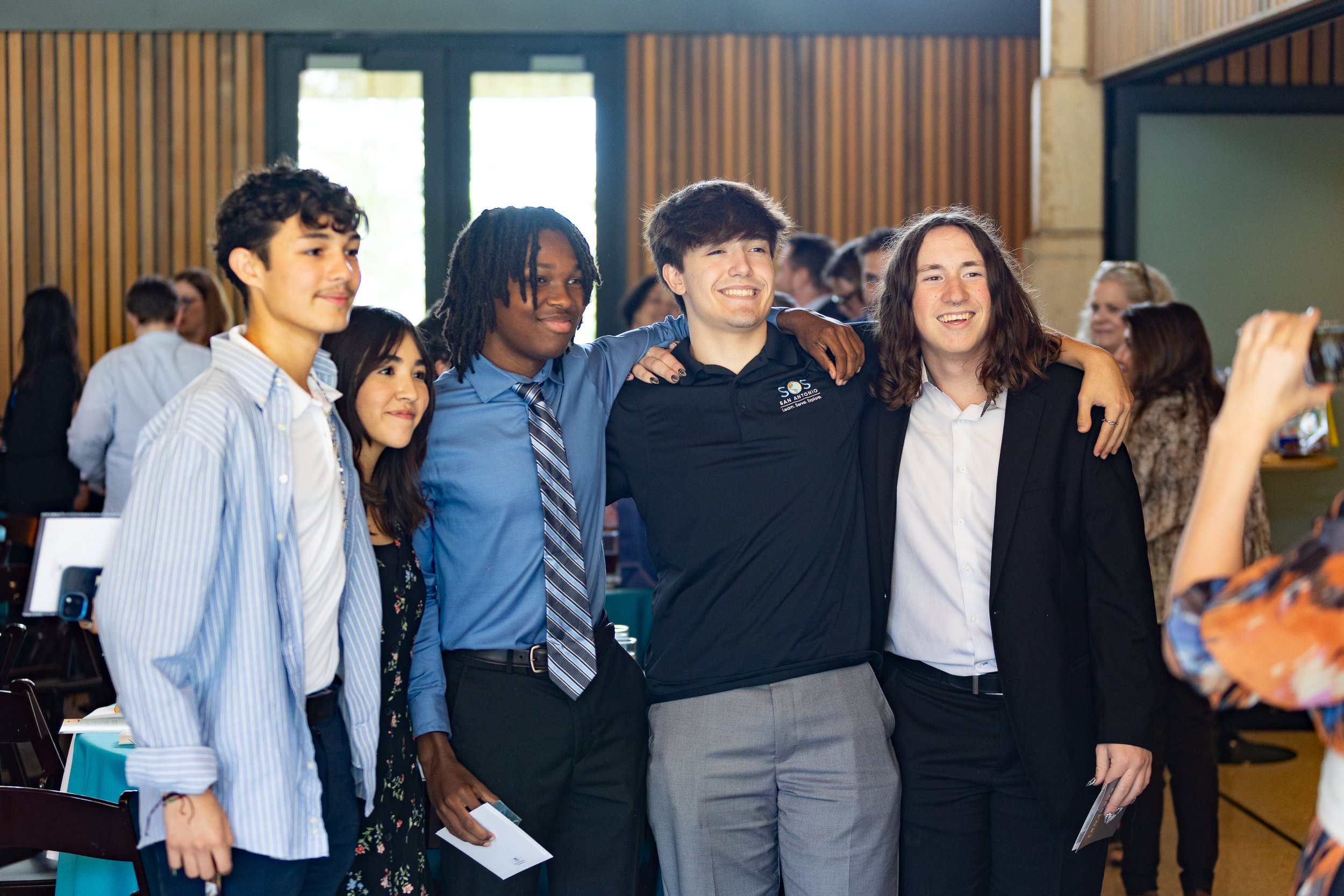 Group of five young diverse adults posing with arms around each other, smiling, in a classroom or conference room setting.