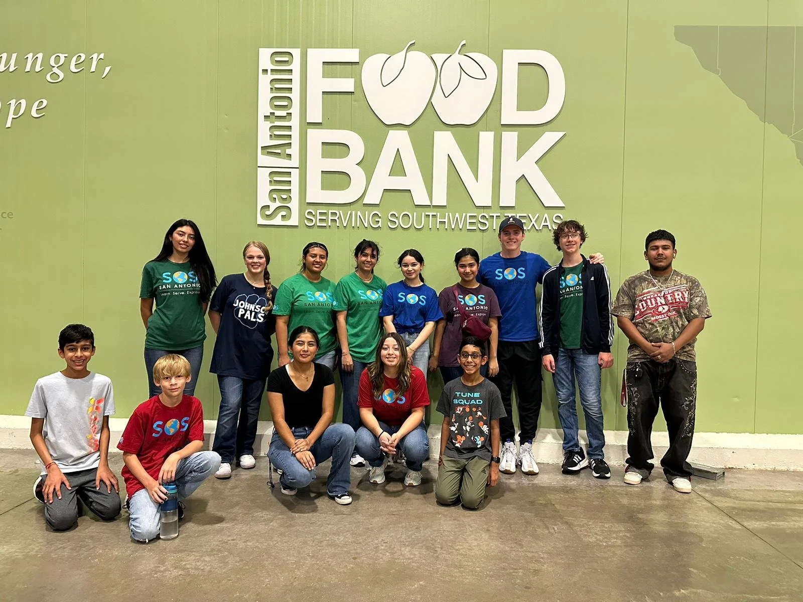 A group of young people posing in front of a large green wall with the logo of San Antonio Food Bank, including some wearing shirts with the food bank logo and others in casual clothing. The group is smiling and standing or sitting for the photo.