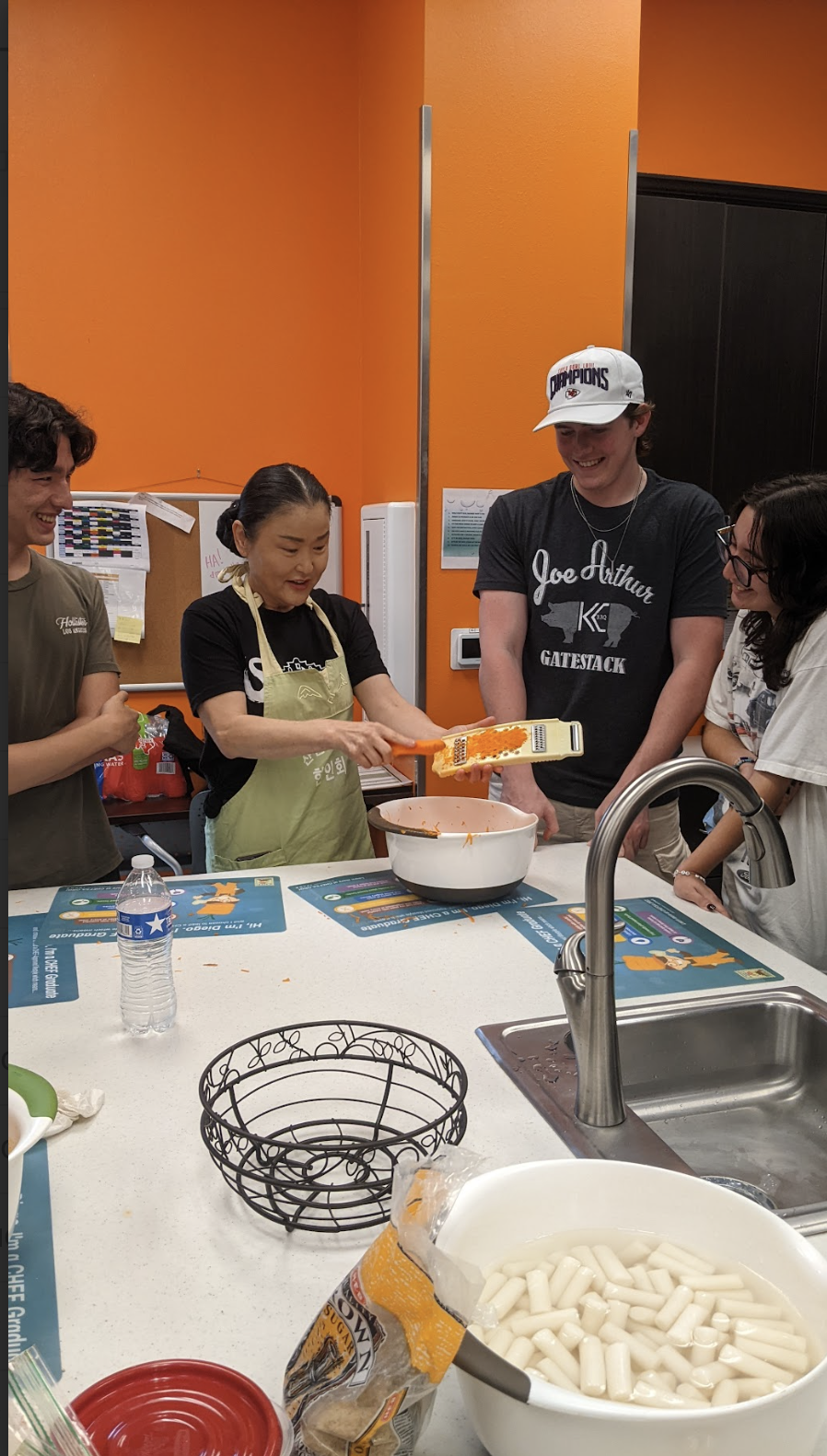 A woman grating orange cheese into a bowl while three young people watch and smile in a brightly lit kitchen with orange walls.