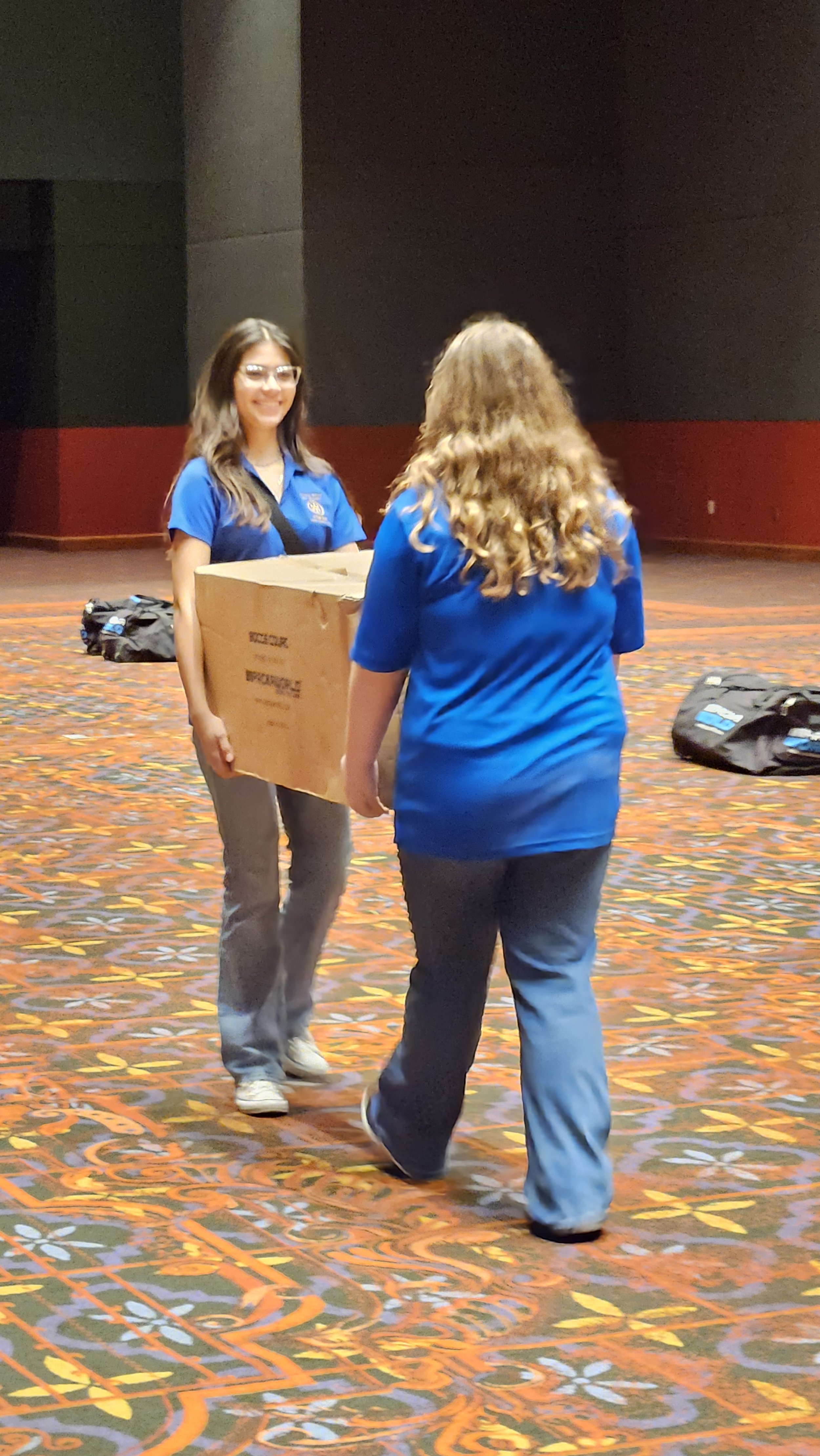 Two women in blue shirts exchanging a large cardboard box in a spacious indoor setting with a colorful patterned carpet.