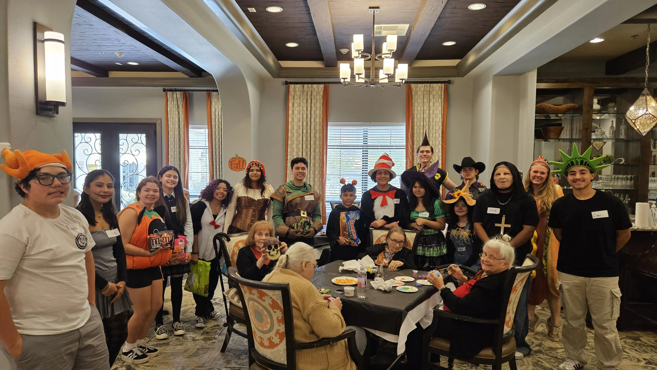 Group of people dressed in costumes, including pirates, witches, and superheroes, gathered around elderly women at a Halloween party in a decorated dining room.
