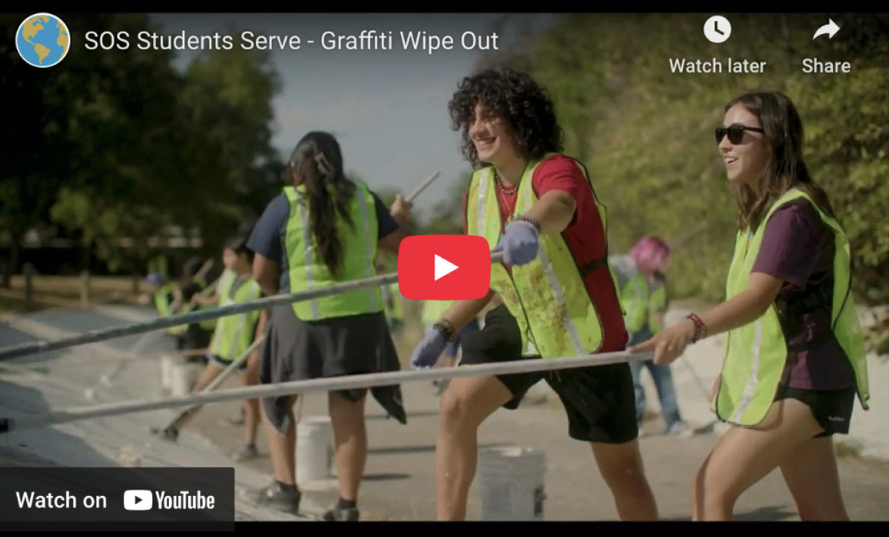 Group of young volunteers in yellow safety vests cleaning graffiti off a surface outdoors on a sunny day.