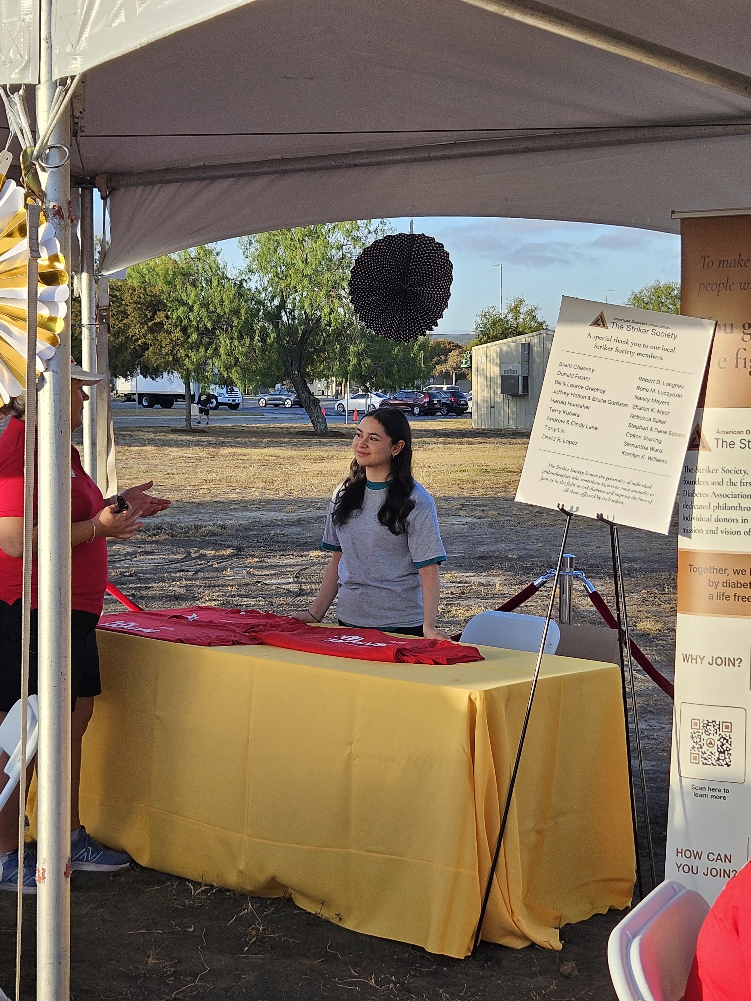 A young woman standing behind a table at an outdoor event, talking to a person in a red shirt. The table is covered with a yellow tablecloth with folded red shirts on top. There are informational posters and a QR code stand next to the table. The bac