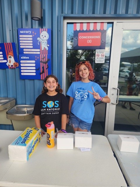 Two young girls standing behind a table, smiling in front of a blue building with a sign for concessions and a menu board.