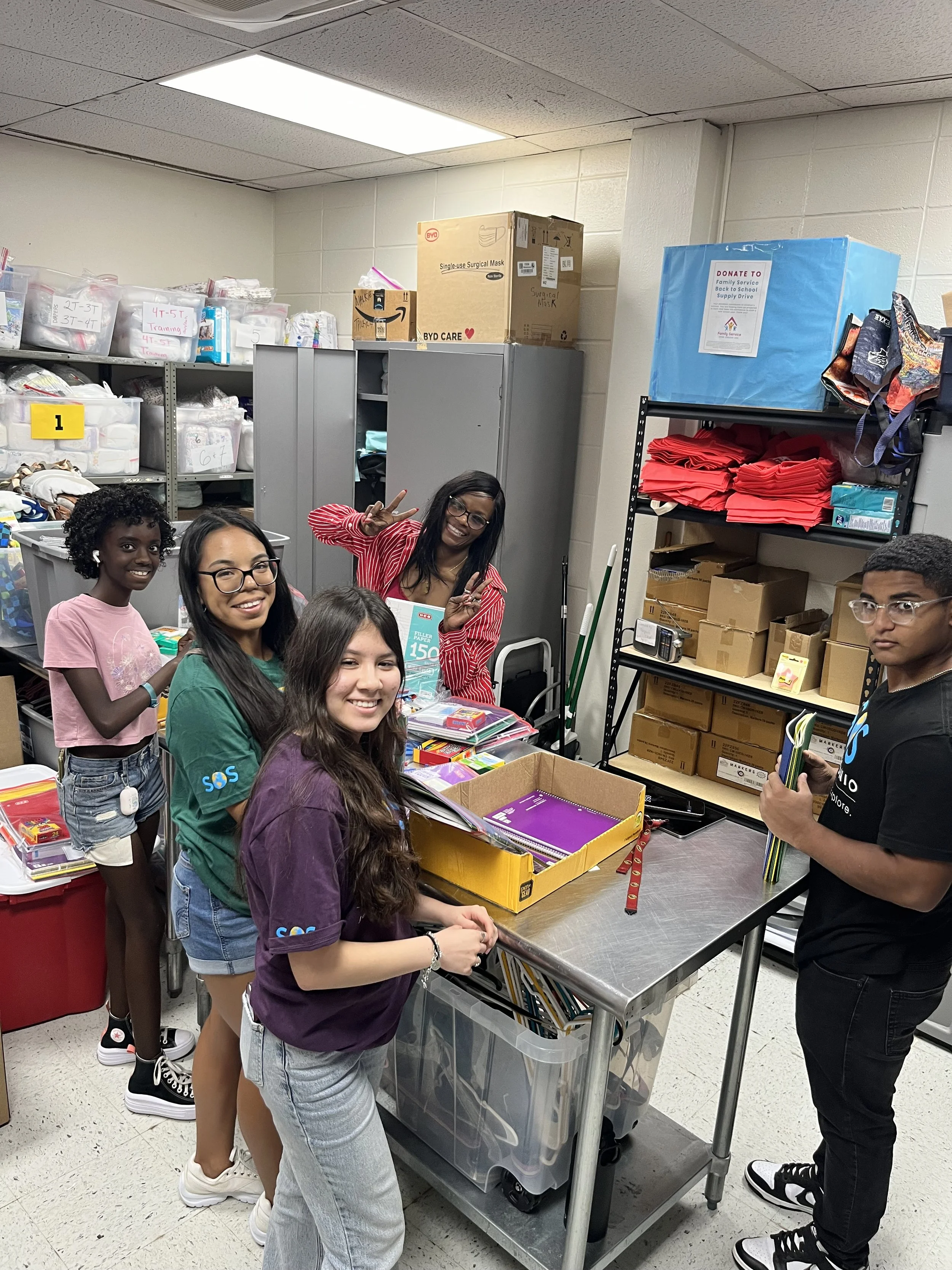 Group of five young people in a storage room, standing around a table with school supplies, smiling and making peace signs.
