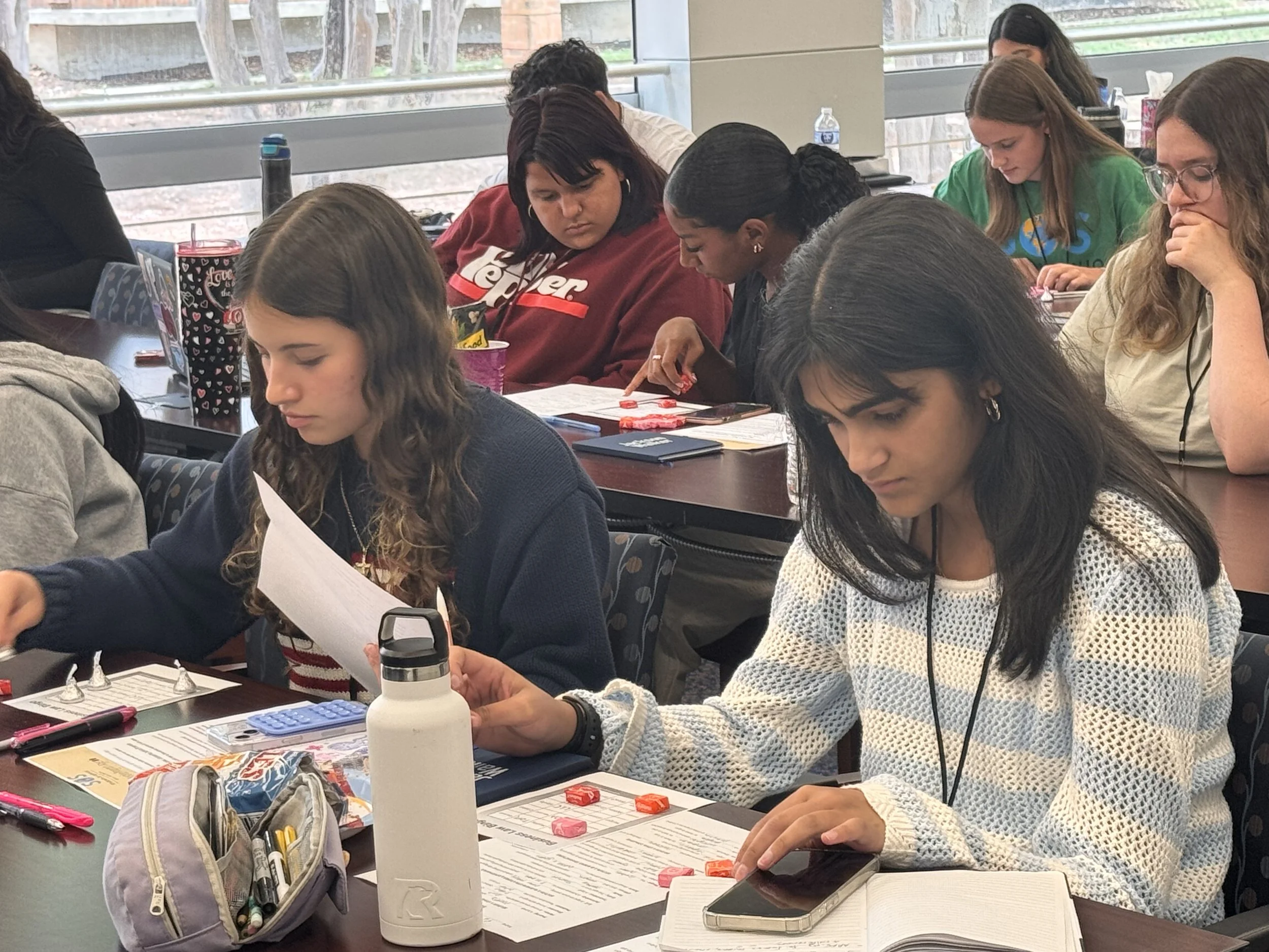 Students sitting at desks in a classroom, working on assignments and reading, with supplies like notebooks, pens, and water bottles visible on the tables.