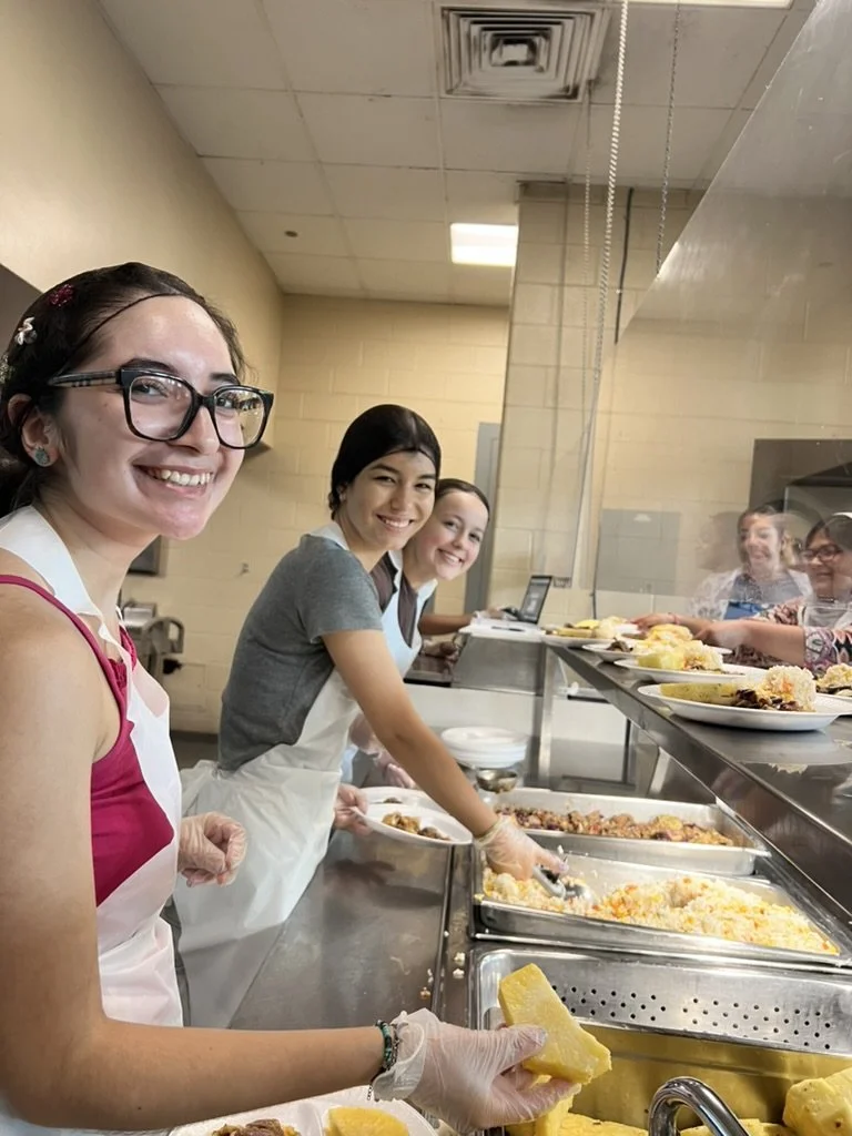 Four smiling women working at a food service counter with trays of food, some wearing aprons and gloves, in a cafeteria or cafeteria-style setting.