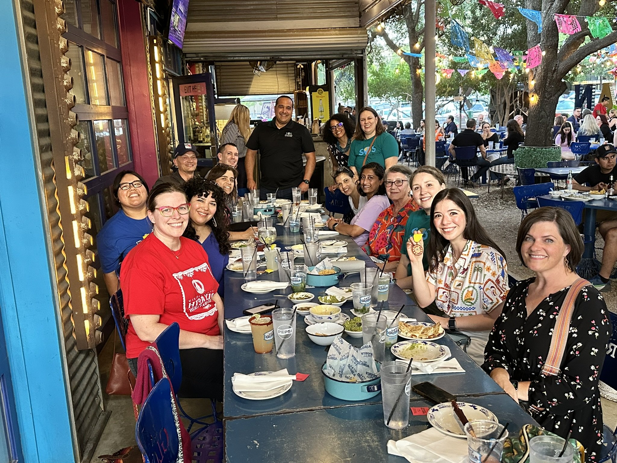 A group of people sitting and standing around a long table at an outdoor restaurant, smiling for the camera. The table has plates of food and drinks, and colorful banners are hanging from trees in the background.