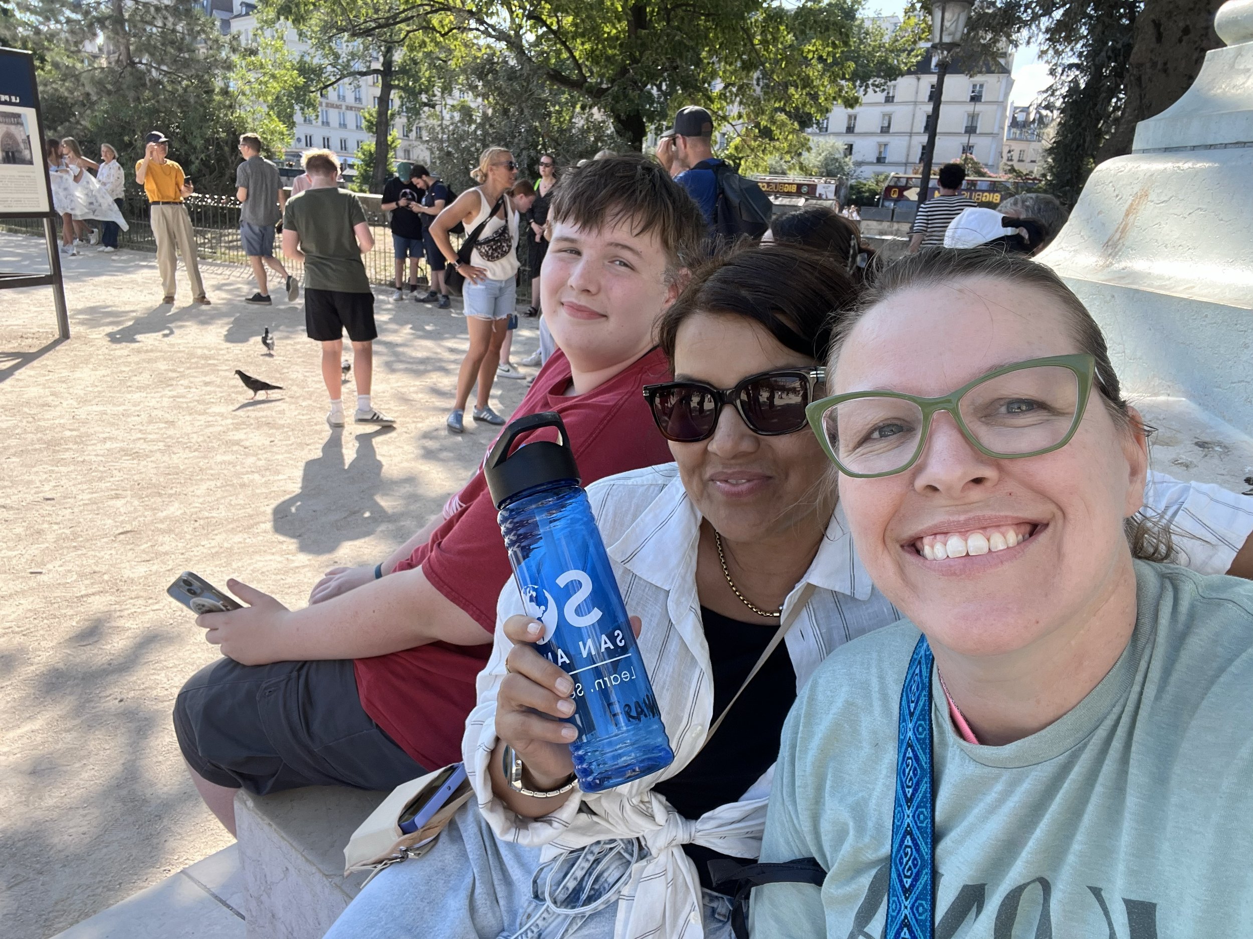 Three smiling people sitting outdoors in a park, with a group of people in the background and trees, holding water bottles and taking selfies.