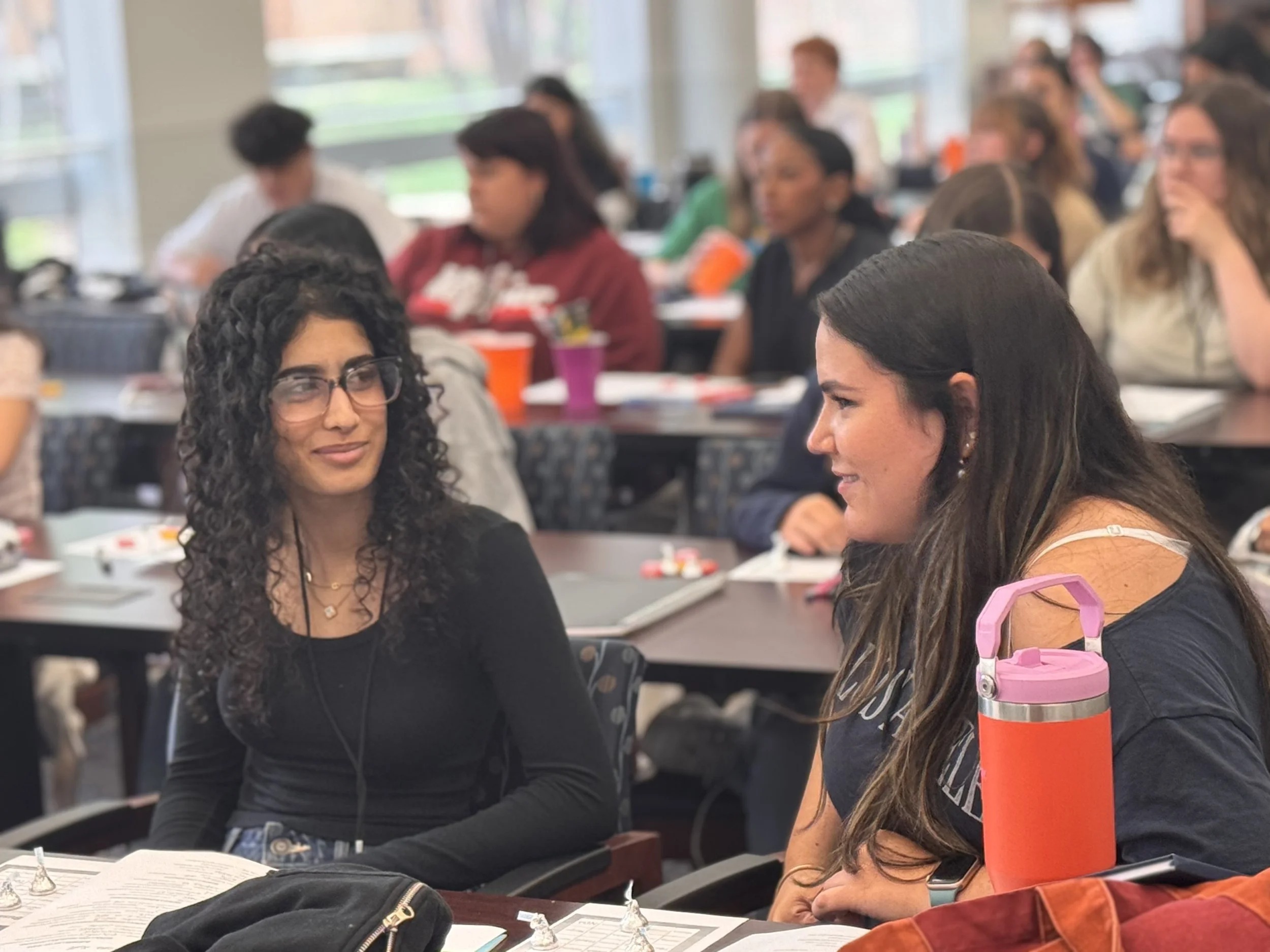 Two women engaged in conversation in a crowded classroom or conference setting, with many other people seated at tables in the background.