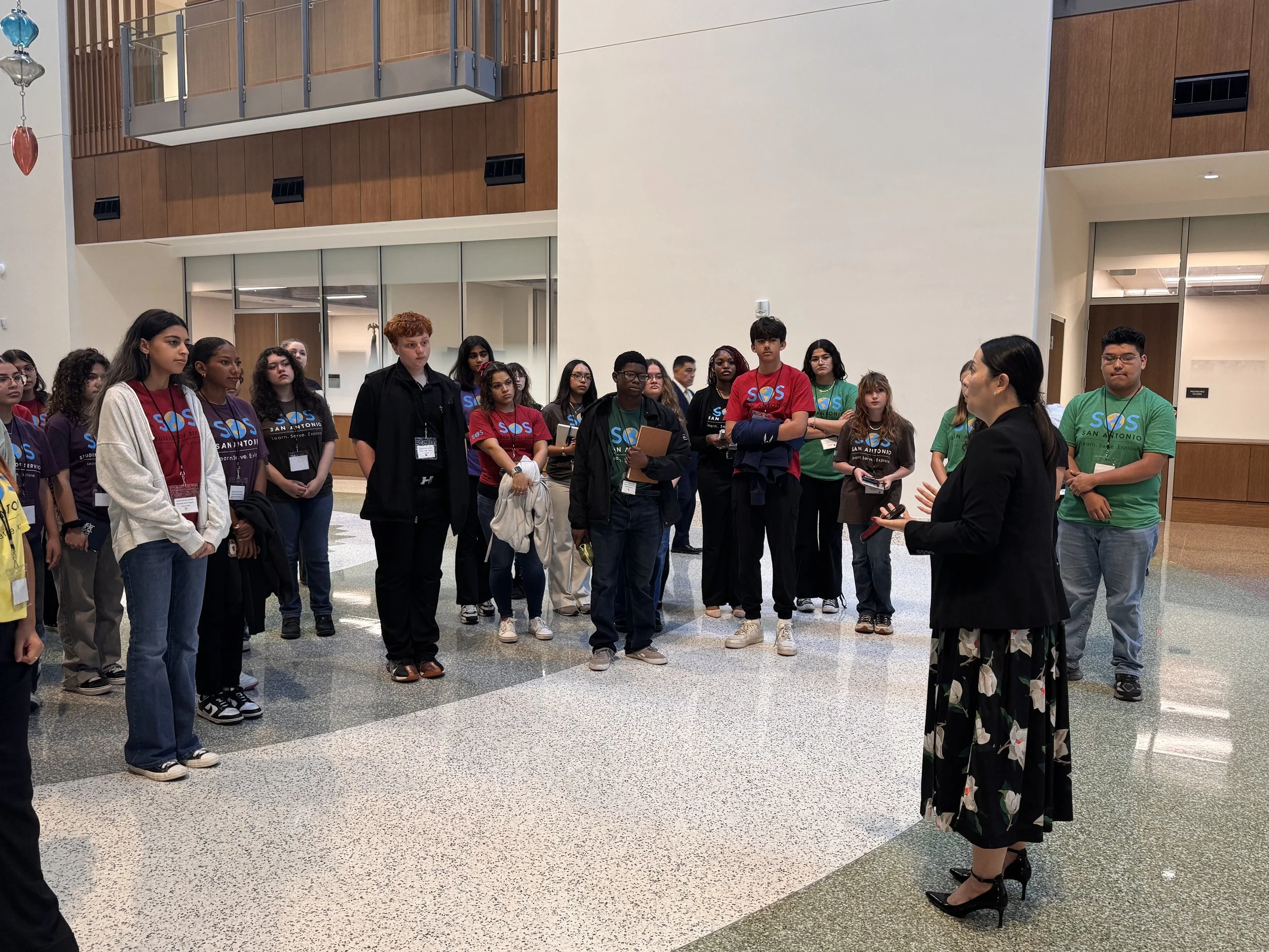 A group of diverse young students listening to a woman speaking in an indoor space with modern architecture, large windows, and a netted balcony.