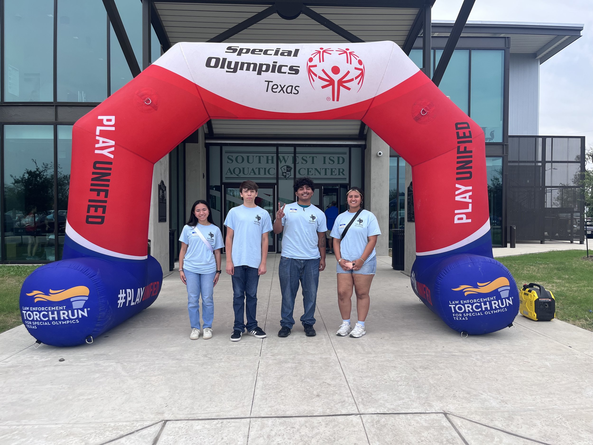 Four people standing under an archway with the words 'Special Olympics Texas' and '#PLAYUNIFIED' at the South West ISD Aquatic Center. The archway is branded with the Law Enforcement Torch Run for Special Olympics Texas.