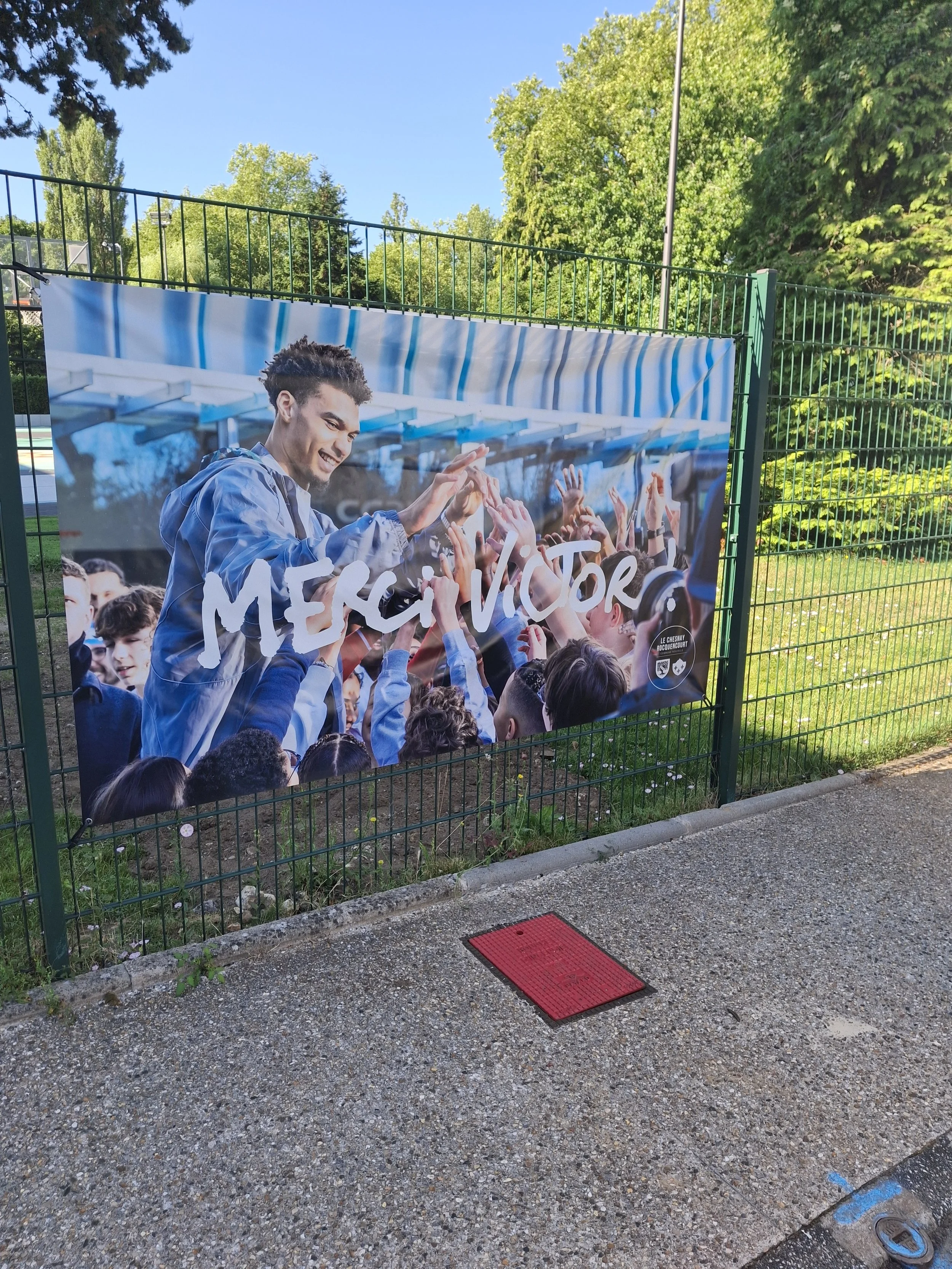 A large banner showing a young man in a blue jacket greeting and high-fiving children in a schoolyard. The words 'MERCY VICTOR' are written across the banner.