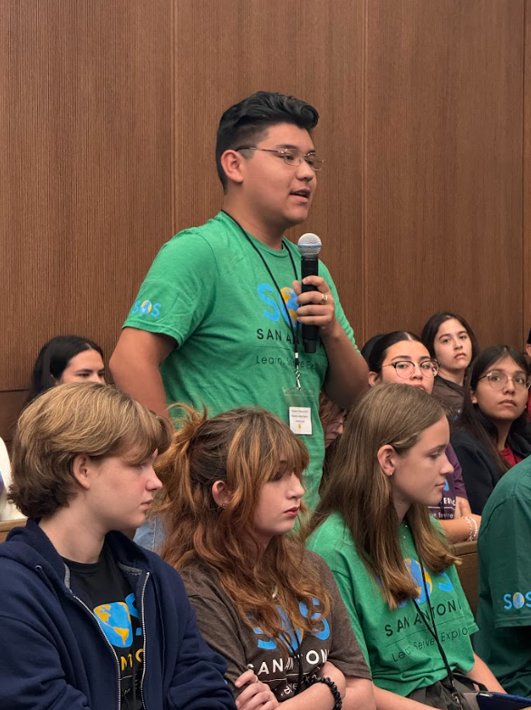 Young man standing in a room with a microphone, speaking to an audience of young students seated and listening attentively, all wearing matching T-shirts with the SOS San Diego logo.