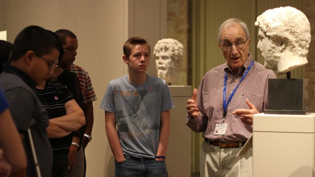 An older man with glasses and a checkered shirt giving a presentation about sculptures to a group of young boys and men at a museum.