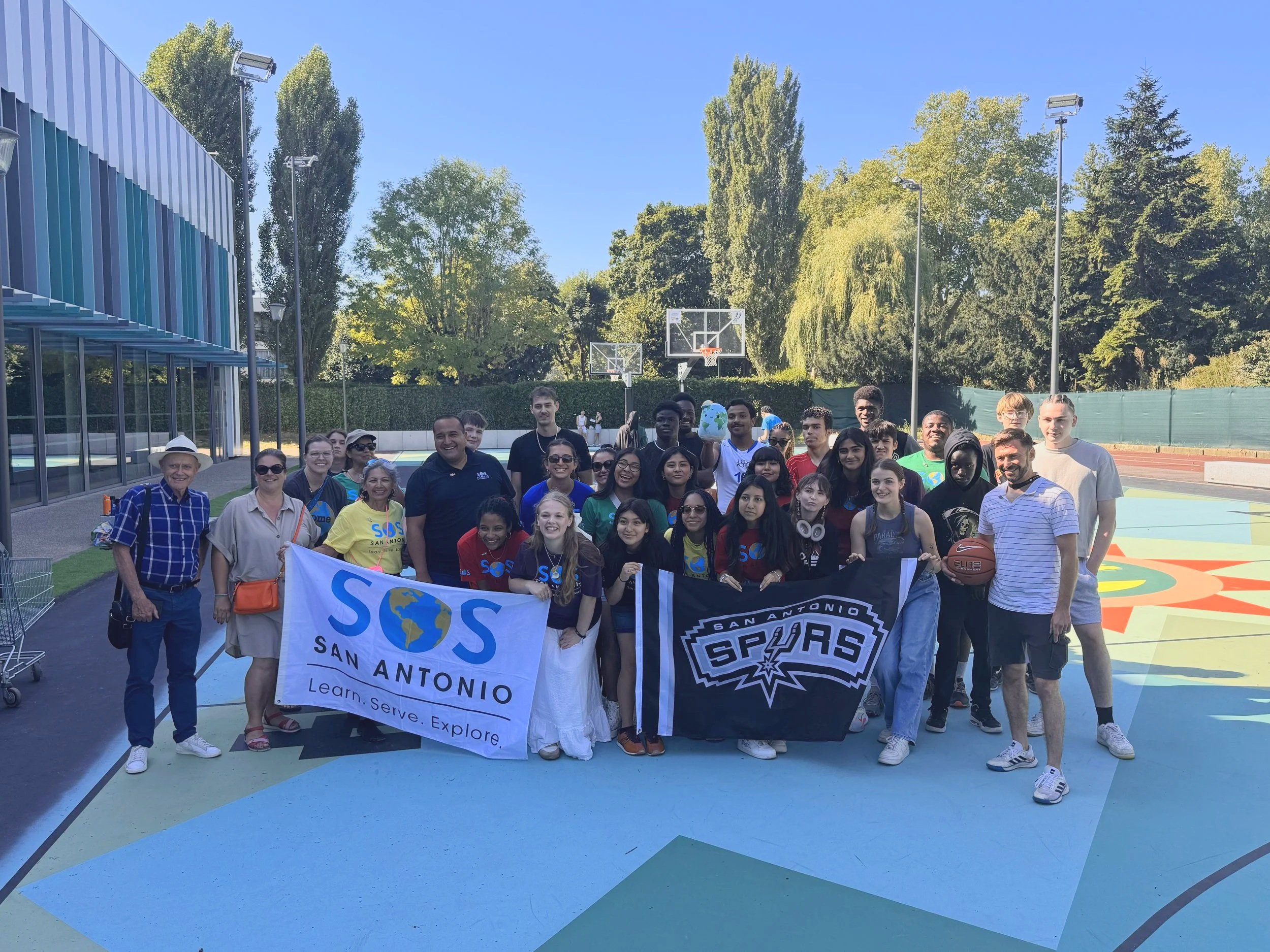 Group of diverse young people and adults on an outdoor basketball court holding banners for San Antonio. Several individuals are smiling, some wearing sunglasses, and one person is holding a basketball. The background features tall trees, a clear blue sky, and nearby sports equipment.