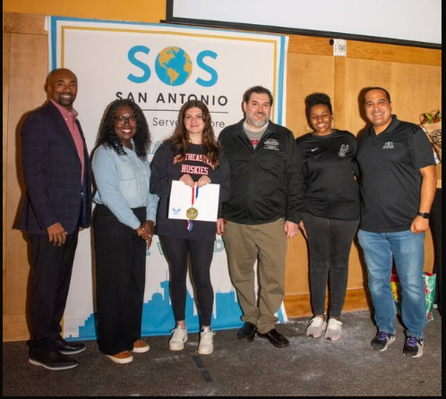 Group of six people standing together at an event, with a banner behind them reading "SOS San Antonio." One woman is holding a certificate, and they are smiling for the photo.