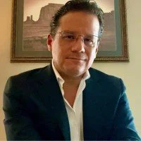 A man with dark hair and glasses, wearing a dark blazer and white shirt, sitting indoors with a framed picture on the wall behind him.