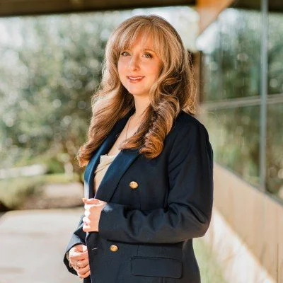 A young woman with long wavy blonde hair smiling, wearing a dark blazer over a white top, standing outdoors near a building with glass windows.
