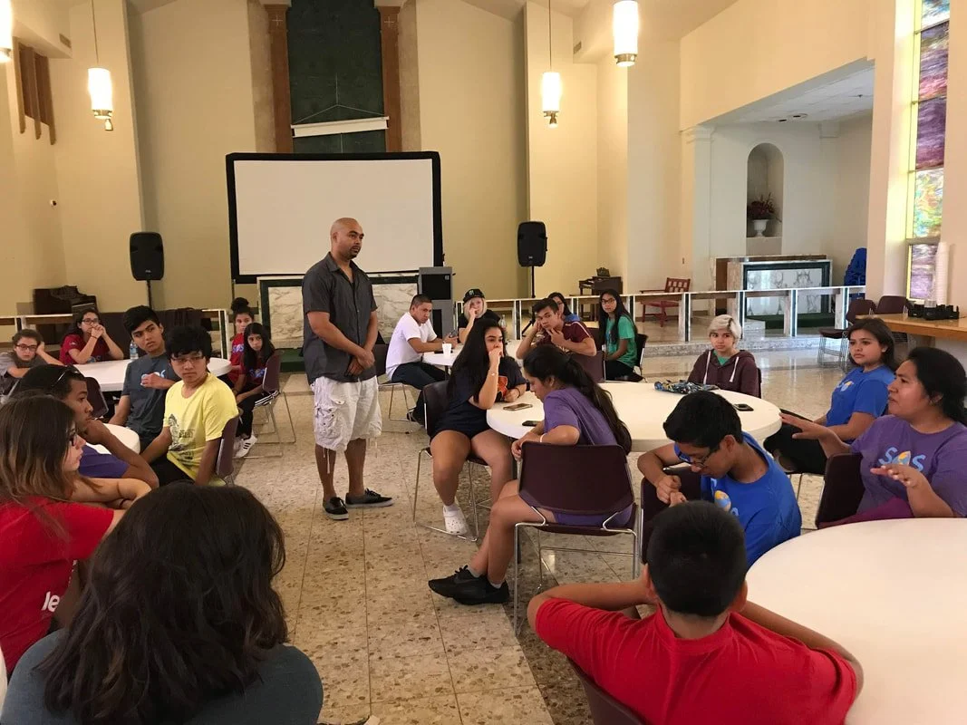 A group of teenagers sitting at round tables in a spacious room with a speaker standing in the middle. There is a white projection screen and speakers in the background. The room has high ceilings and soft lighting.