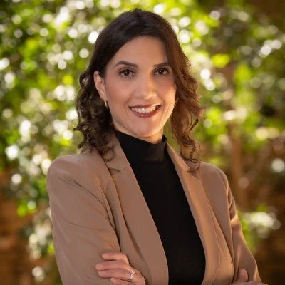 A woman with shoulder-length dark hair, wearing a beige blazer over a black top, smiling with arms crossed, standing outdoors with greenery in the background.