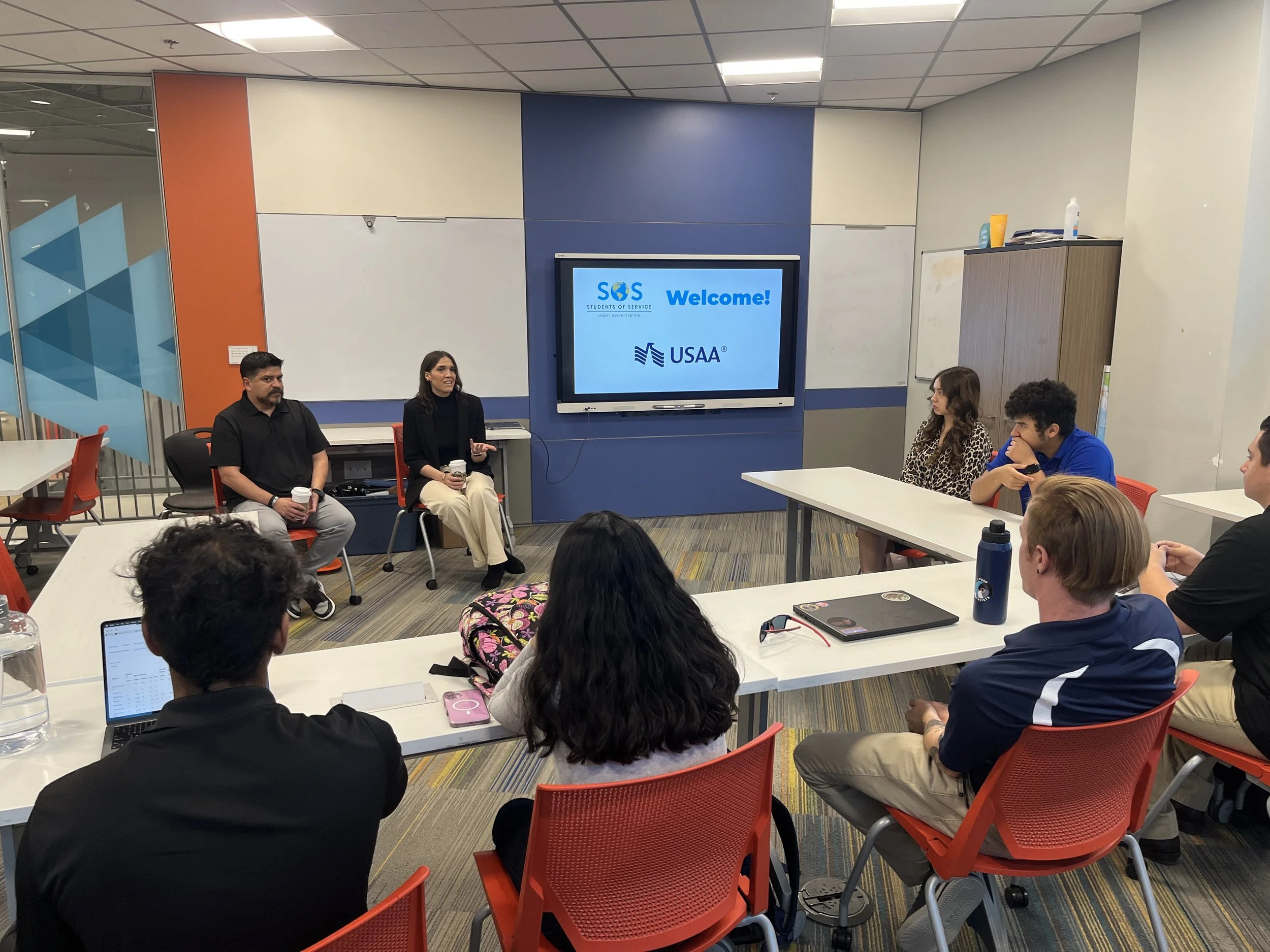 A group of nine people in a classroom or conference room, listening to two women speaking at the front. The room has a large screen displaying the words 'SOS Welcome! USA' and an orange and blue color theme. The attendees are seated at tables, some with laptops, notebooks, and drinks.
