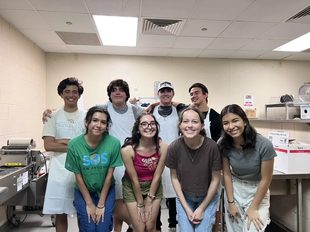Group of nine young people smiling and posing in a kitchen or food preparation area, some wearing aprons.