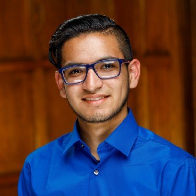 A young man with dark hair, glasses, and a light beard, wearing a blue dress shirt, smiling in front of a wooden background.