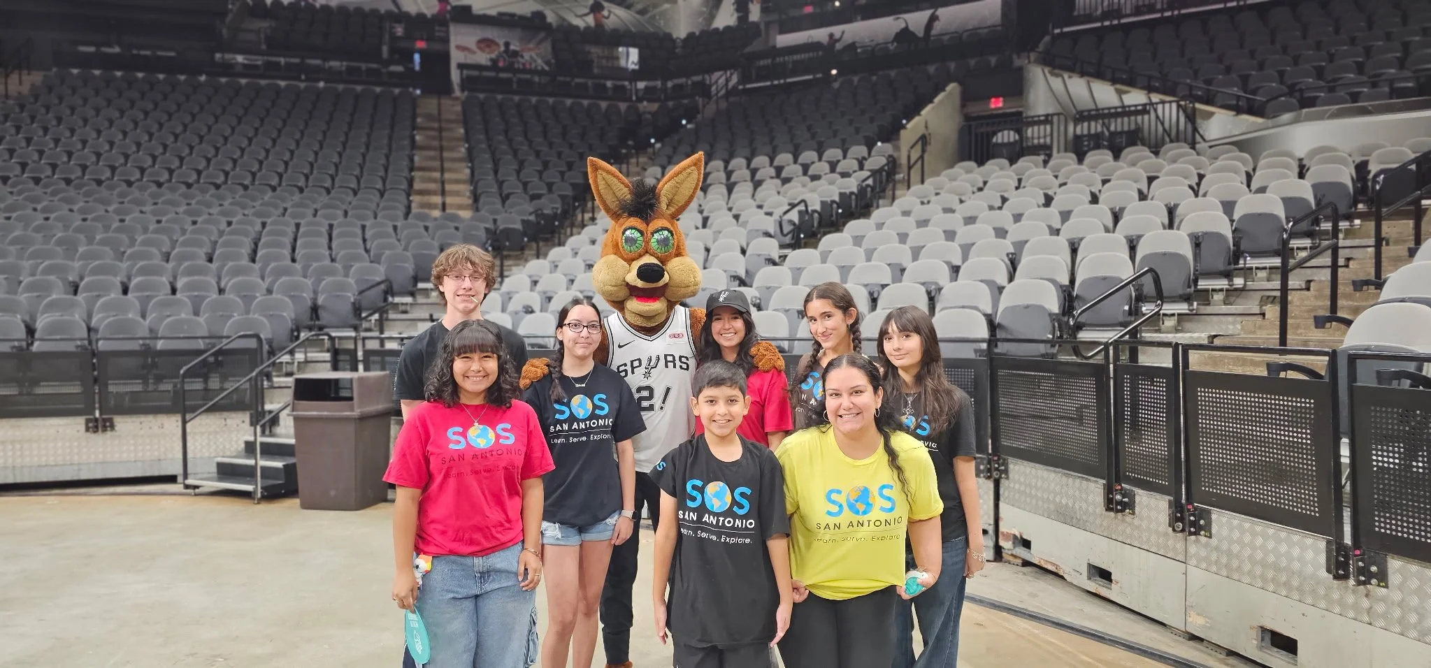 Group of seven kids and a person in a dog mascot costume standing inside an empty arena, smiling for a photo.